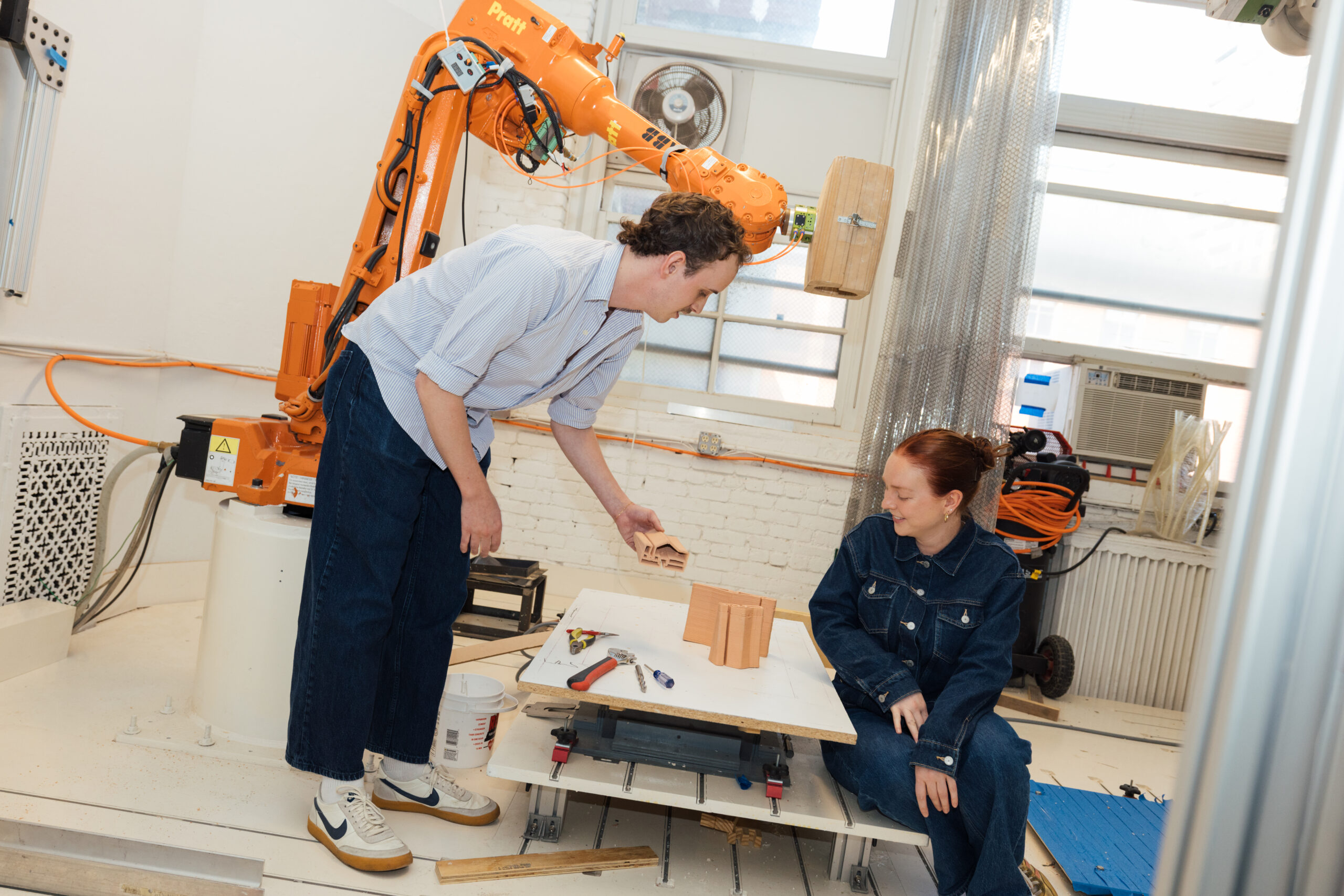 Two people work together in a fabrication studio beside an industrial robotic arm. One stands and holds a small architectural model component while speaking to the other, who sits next to a low platform table with tools and terracotta-colored printed forms. The workspace includes exposed brick walls, windows, equipment, and coiled cables.