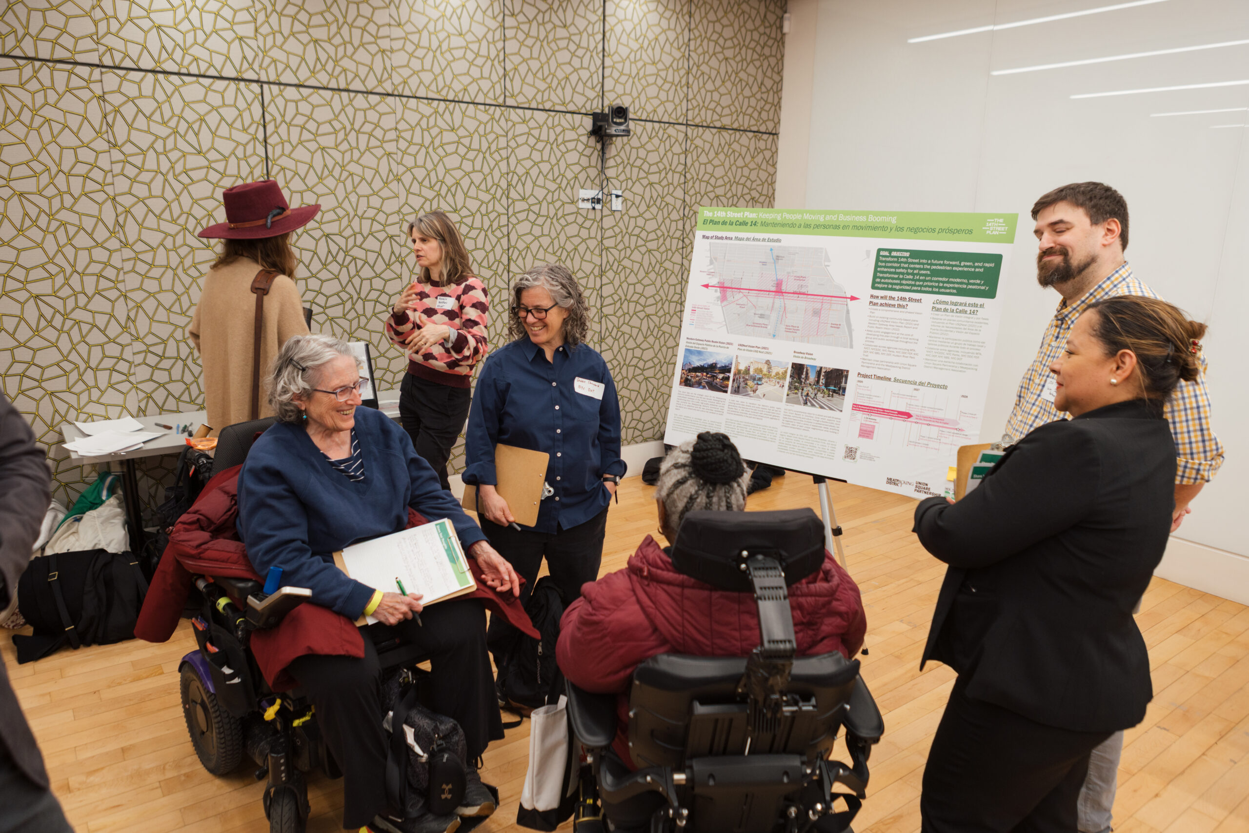 A group of people engaged in discussion at a community event. Two women, one in a wheelchair and the other seated beside her, smile and interact with each other. Another woman gestures while speaking to a man, who is listening attentively. A poster titled "The 14th Street Plan: Keeping People Moving and Business Booming" is displayed nearby. The background features a patterned wall, and various papers are spread across a table.
