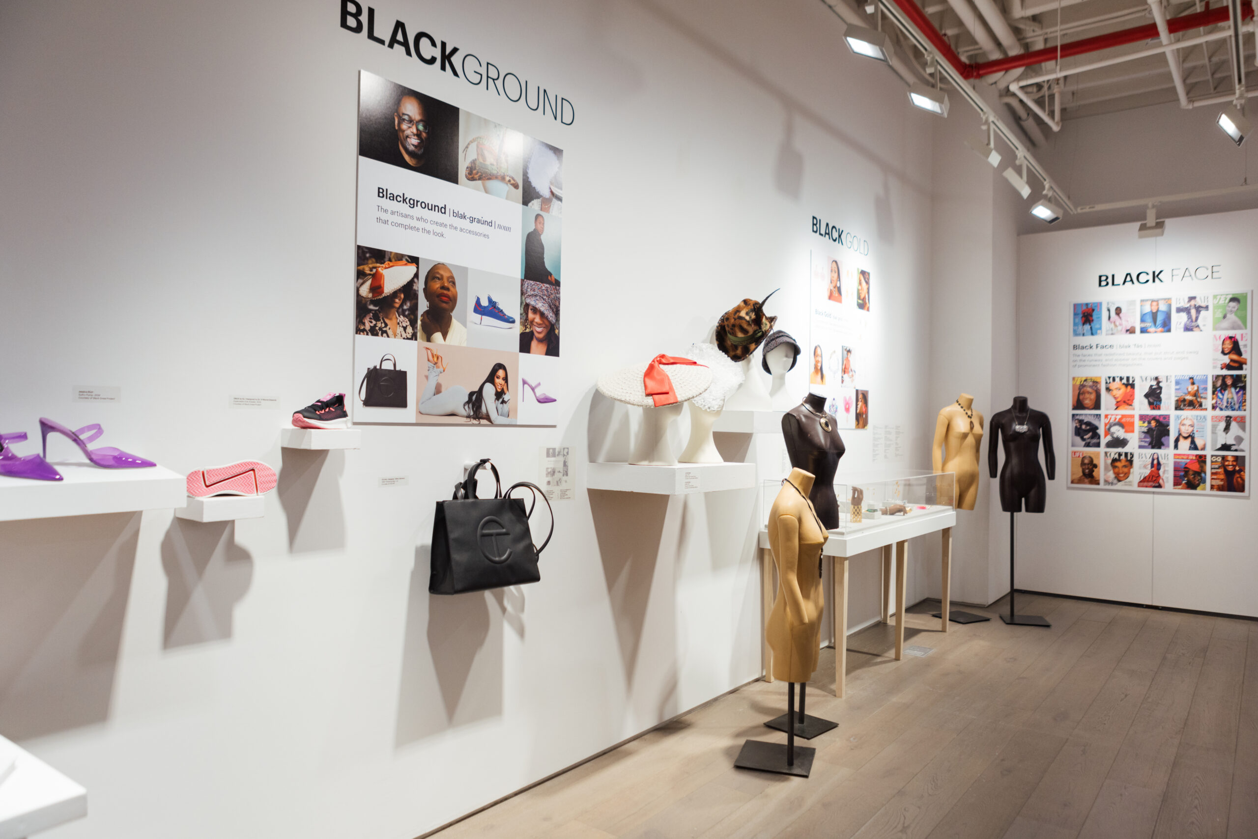 An exhibition space with white walls showcases various fashion accessories. On the left side, there are shelves displaying purple heels, pink sneakers, and a black handbag. Mannequins dressed in brown and black outfits stand in the foreground, while descriptive panels titled "BLACKGROUND," "BLACK GOLD," and "BLACK FACE" highlight featured artists and their contributions. The floor is wooden, and the space is well-lit with overhead lights.