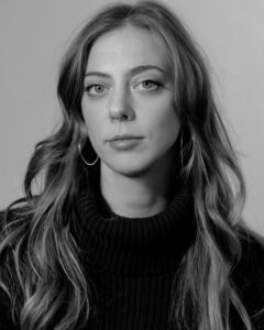 Black-and-white portrait of a woman with long, wavy hair wearing a dark turtleneck sweater. She faces the camera with a neutral expression. She has a small hoop nose ring and large hoop earrings. The background is plain and softly lit.