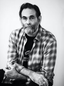 Black-and-white portrait of a bearded man seated with his arms loosely crossed. He wears a plaid button-down shirt over a dark T-shirt. He looks directly at the camera with a calm, neutral expression. The background is plain and softly lit.
