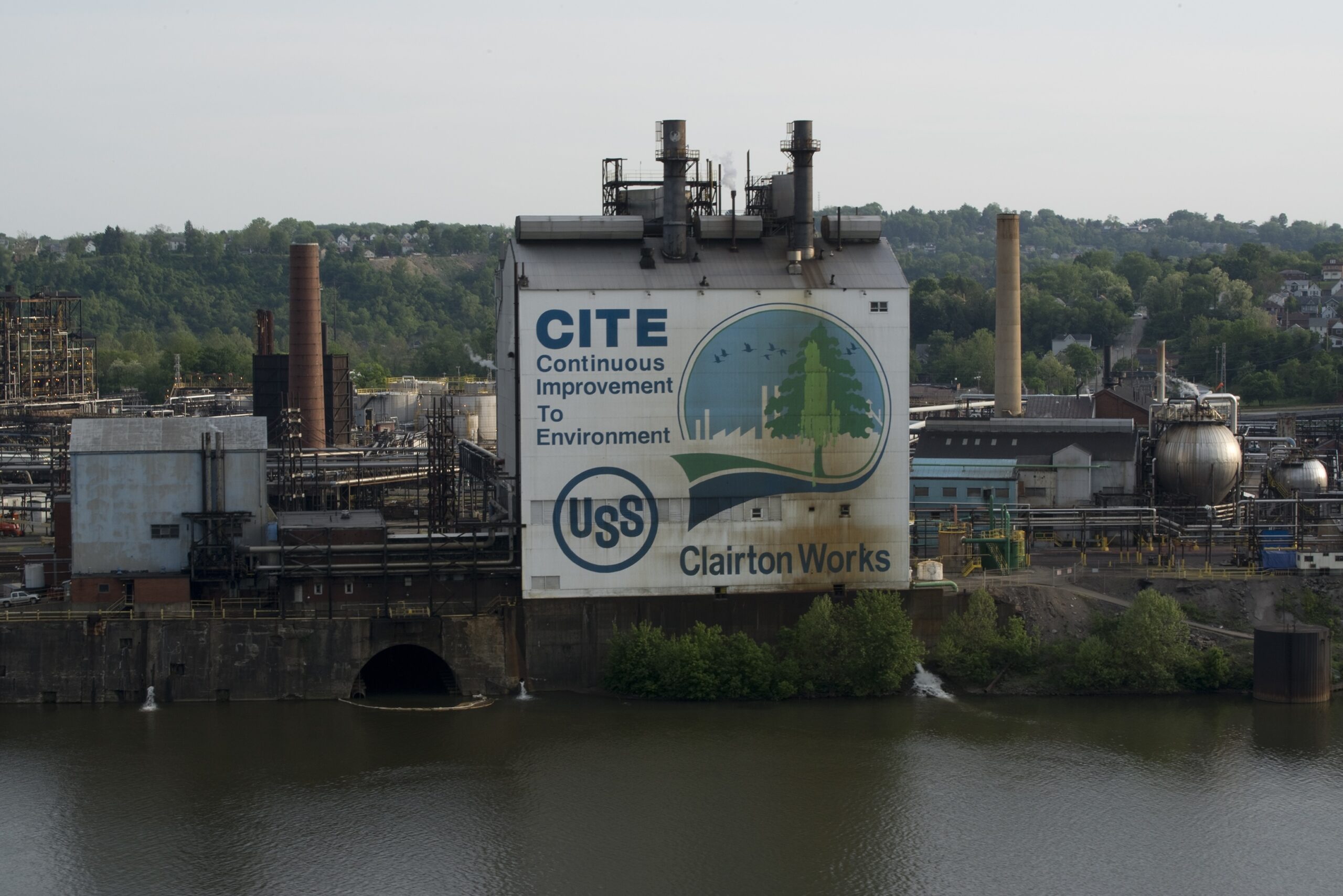 A large industrial building with a prominent mural on its side, featuring the text "CITE Continuous Improvement To Environment" and "USS Clairton Works." The building is situated near a body of water, with various industrial structures and smokestacks visible in the background. Lush green hills and trees are visible beyond the industrial area.