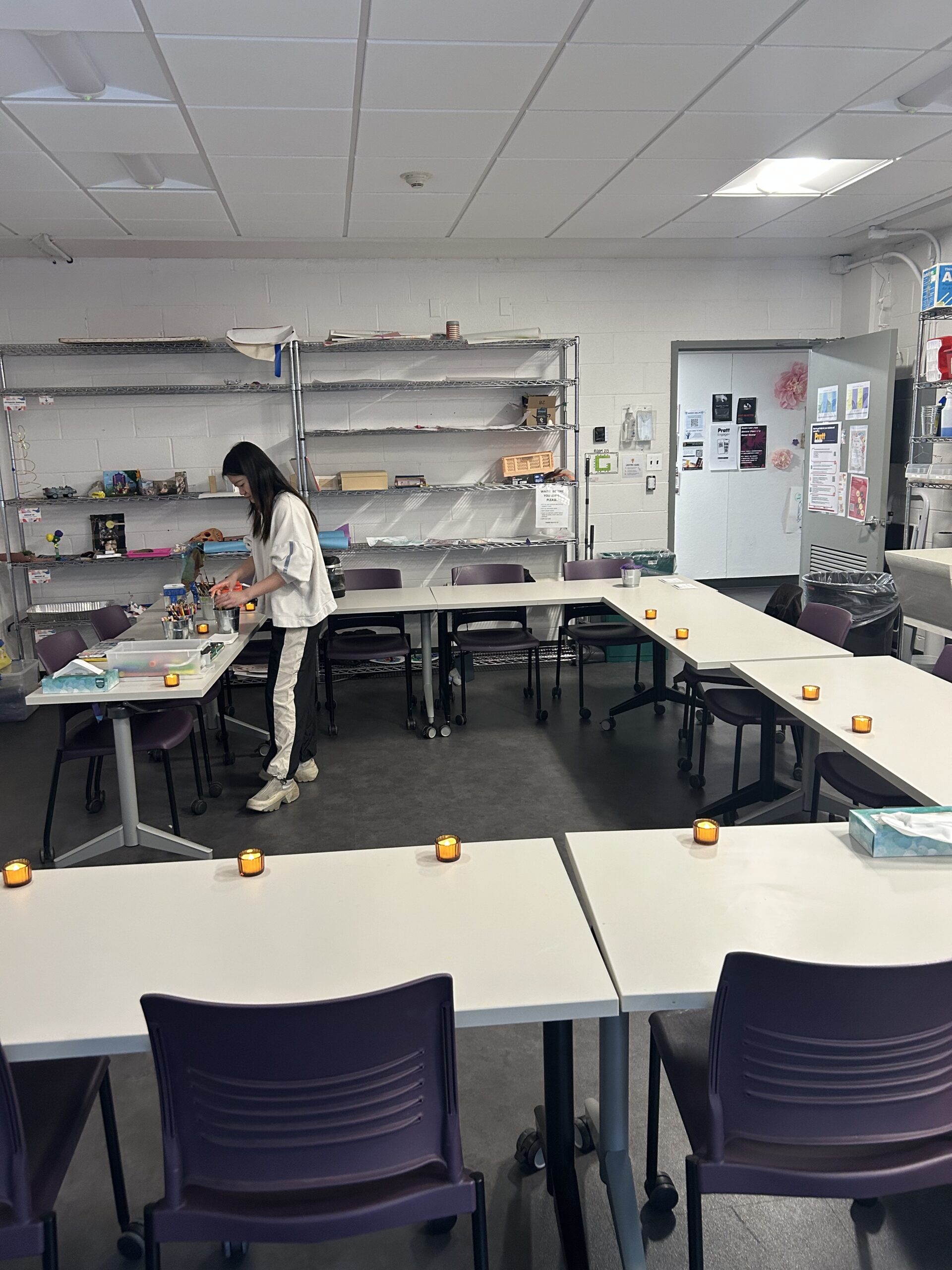 Wide view of a classroom arranged in a U-shape with white tables and purple chairs on casters. Small LED votive candles are spaced evenly along the tables. A woman stands near a supply table, organizing art materials. Metal shelving with craft supplies lines the back wall. An open door reveals posters and notices on the wall. Overhead fluorescent lights illuminate the room.