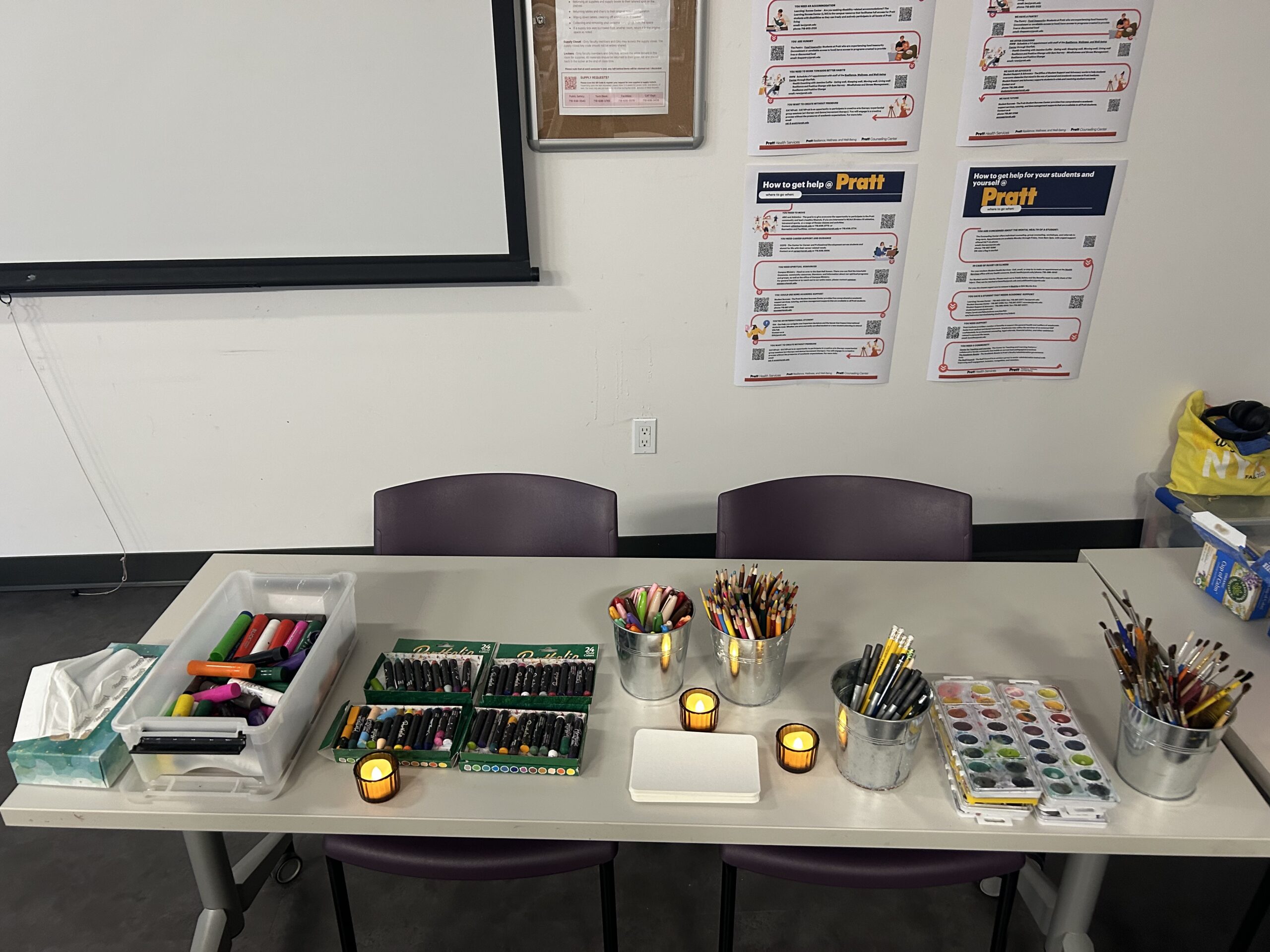 Close view of an art supply table in a classroom. Containers hold colored pencils, markers, pens, and paintbrushes. Open boxes of oil pastels and a watercolor palette are arranged neatly on the table. A tissue box and small LED votive candles are placed among the supplies. Behind the table, two purple chairs sit against a wall with informational posters and a projection screen partially visible above.