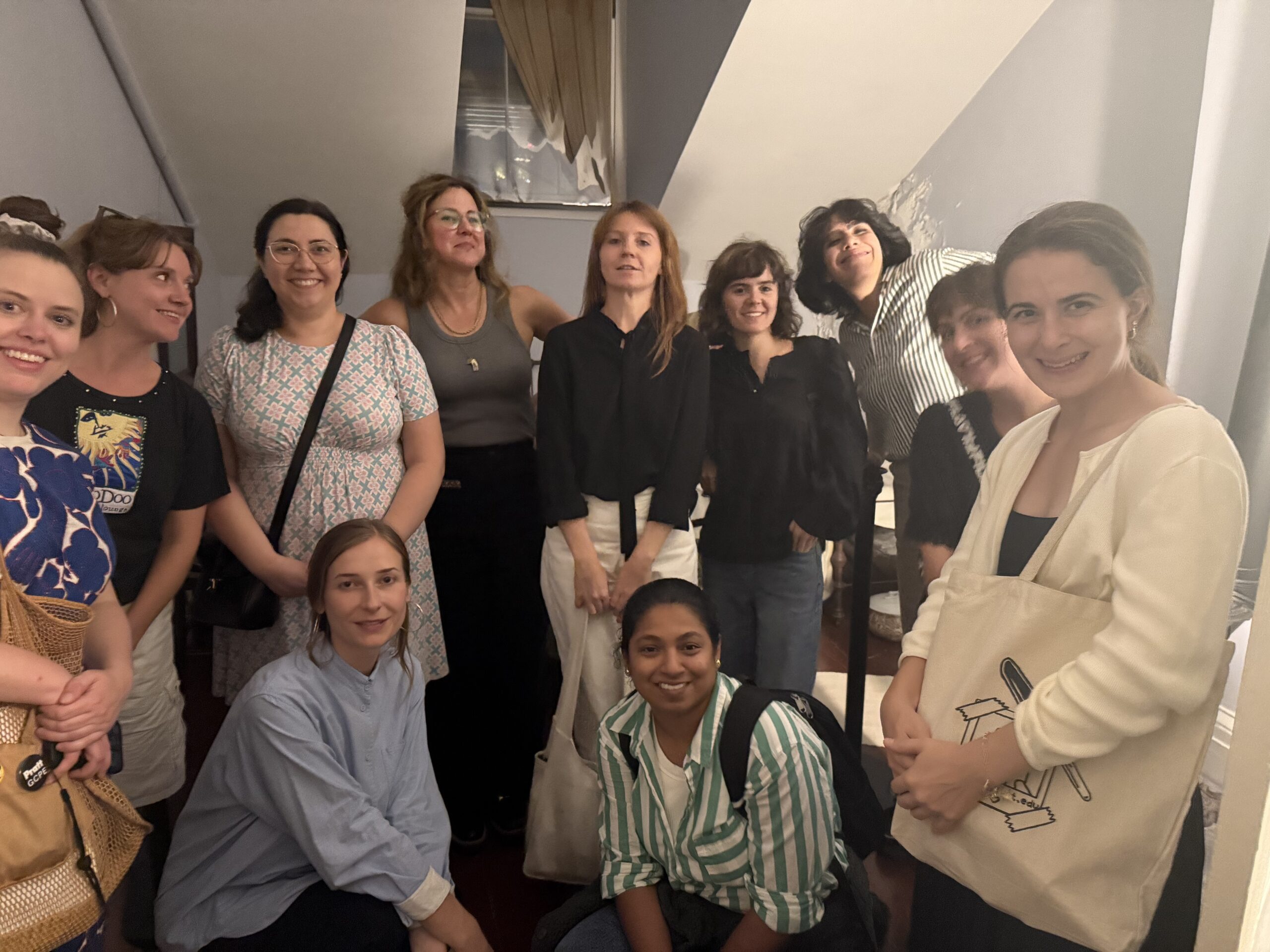 A group of twelve women poses together indoors, smiling and looking towards the camera. They are dressed in a variety of styles, including casual and trendy outfits. The setting appears to be a cozy room with soft lighting and visible architectural details, such as a staircase. Some women are standing, while others are seated, creating a sense of camaraderie and warmth.