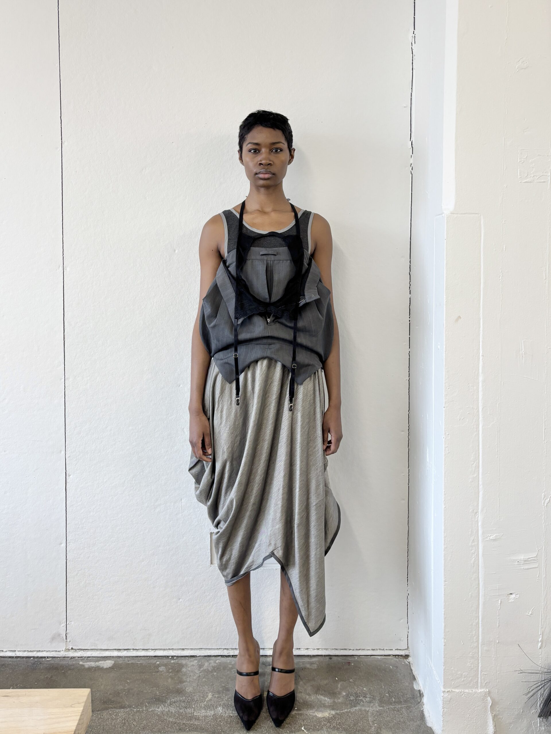 A model stands against a white wall, wearing a layered outfit featuring a sheer black top over a draped gray skirt. The skirt has an asymmetrical hemline and is cinched at the waist with drawstrings. The model wears black high-heeled shoes and has short hair, presenting a neutral expression. The floor is a concrete texture, adding to the minimalist aesthetic of the scene.