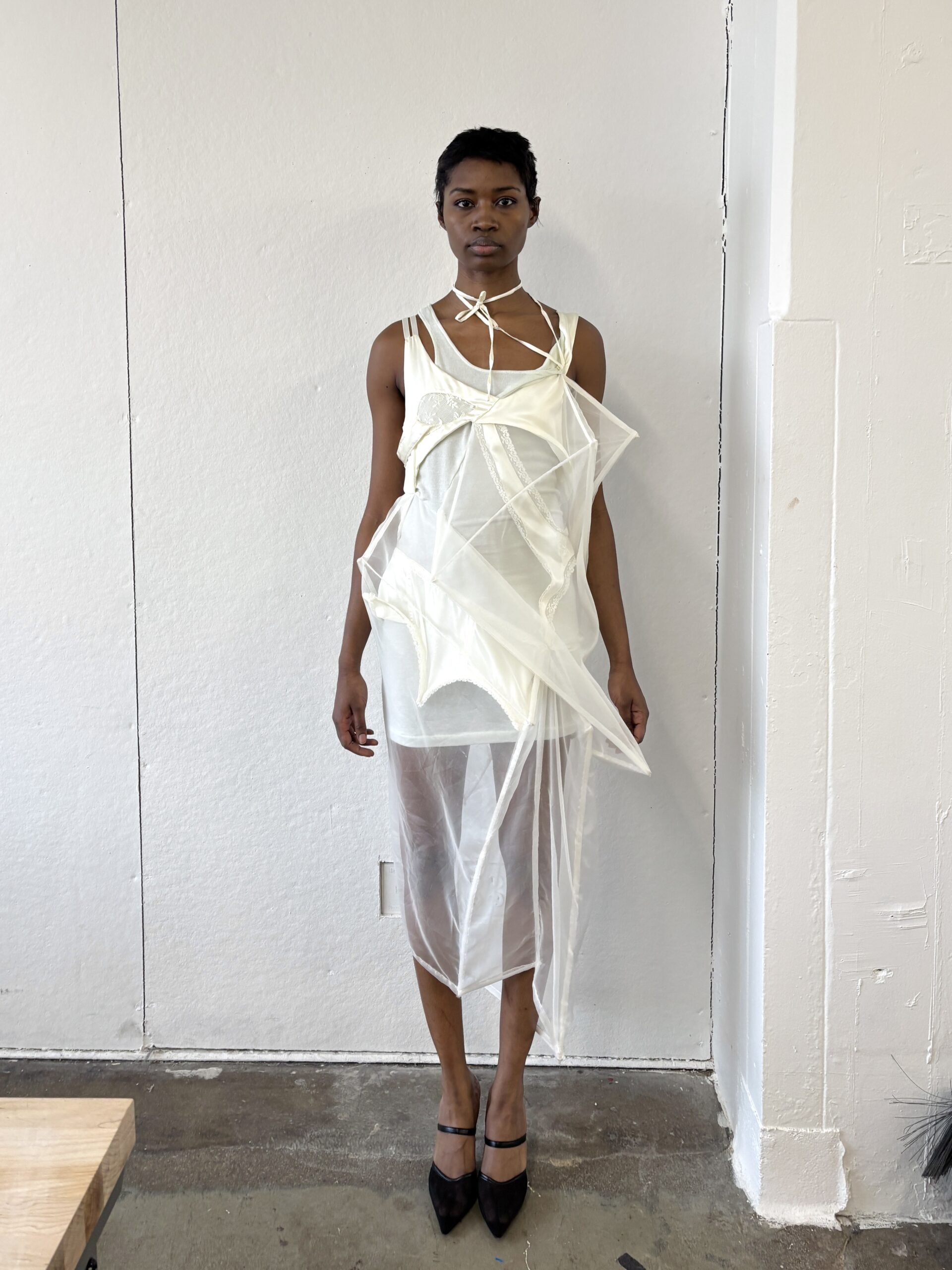 A model stands against a white wall, wearing an avant-garde outfit made of sheer fabrics. The ensemble includes a layered, translucent top with geometric cutouts and a flowing bottom wrap. They have a short hairstyle and are wearing black high-heeled shoes. The setting appears to be a studio with minimalistic decor.