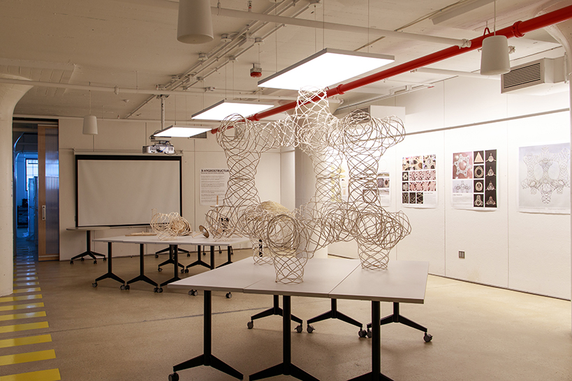 An art installation is displayed in a well-lit gallery space featuring white walls and a polished concrete floor. Several geometric, sculptural forms made of intertwined, light-colored materials are situated on multiple tables. A projection screen is visible on one wall, with dark blue light illuminating the doorway in the background. Various printed artworks are mounted on the walls, displaying intricate designs.