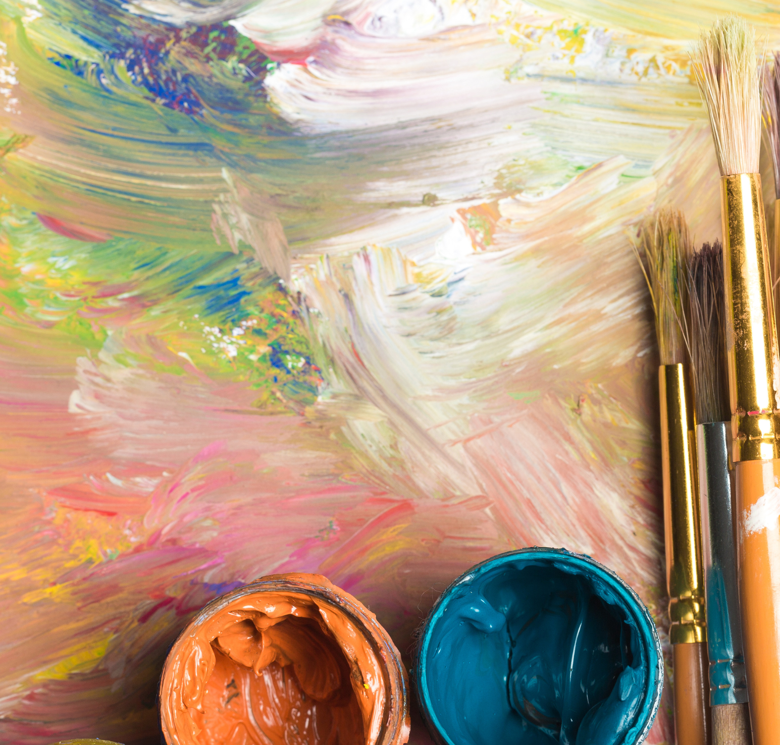 Overhead view of open paint jars filled with thick orange and teal paint resting on a surface covered in multicolored brushstrokes. Additional jars with yellow and red paint are partially visible at the bottom edge. Several paintbrushes with wooden handles and gold metal ferrules lie vertically along the right side of the frame. The background is layered with blended pastel tones, including pink, green, yellow, and white, creating a textured, abstract surface.