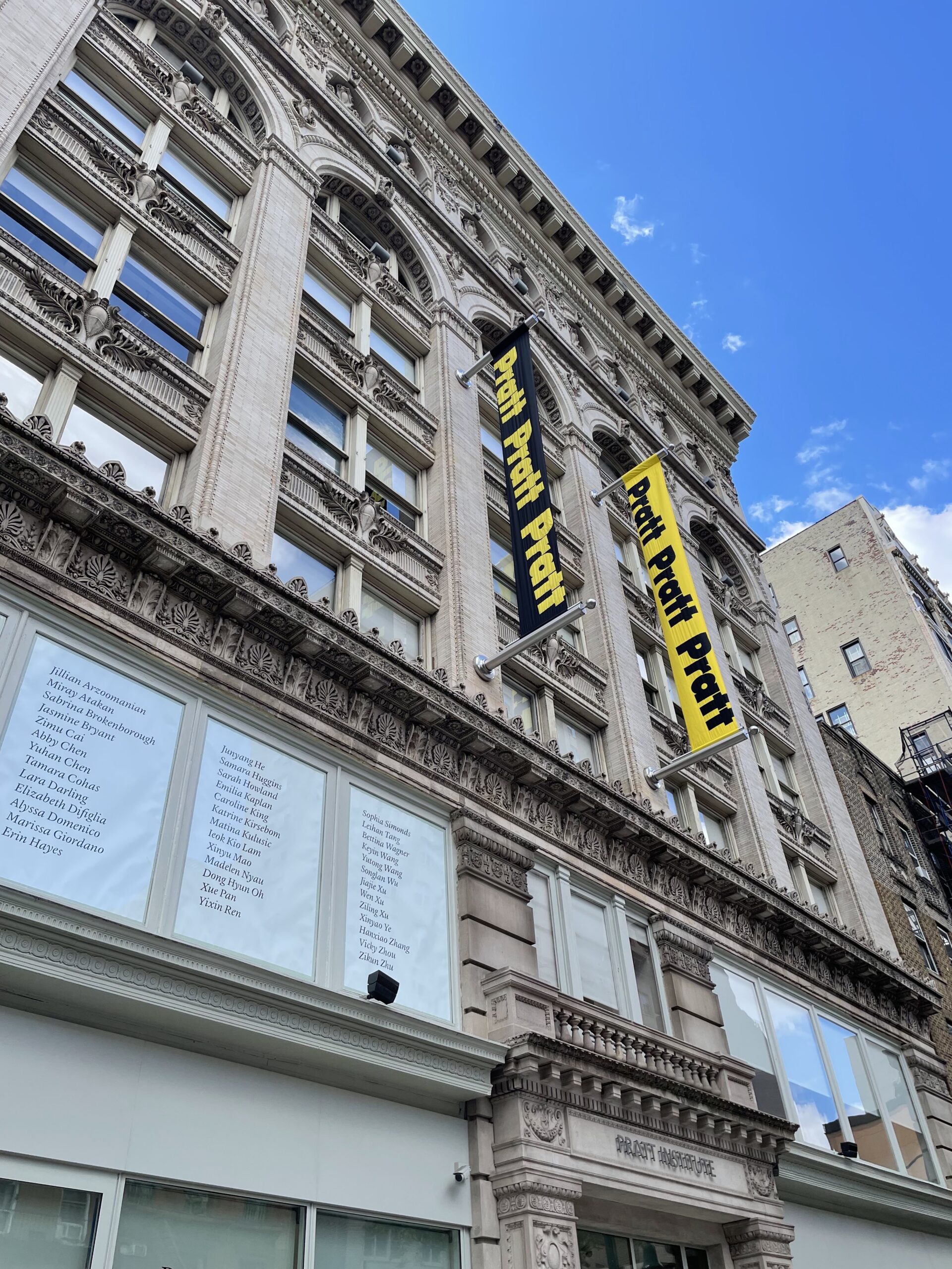 Upward view of an ornate historic stone building with detailed carvings, arched windows, and decorative cornices against a bright blue sky with scattered clouds. Two vertical banners reading “Pratt Pratt Pratt” hang from metal poles—one black with yellow text and one yellow with black text. Large windows on the lower floors display lists of names in black text. The entrance below features carved stonework with “PRATT INSTITUTE” engraved above the doorway.