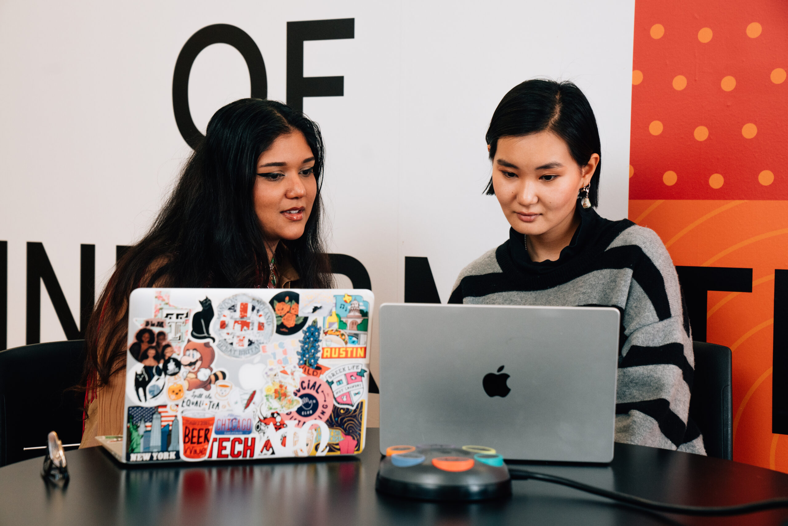 Two young women are seated at a table with laptops in front of them. One woman has long, dark hair and is looking at a laptop covered in colorful stickers, while the other has short black hair and is focused on a silver MacBook. A device with colored buttons is placed on the table in front of them, and a large poster with bold letters is in the background.