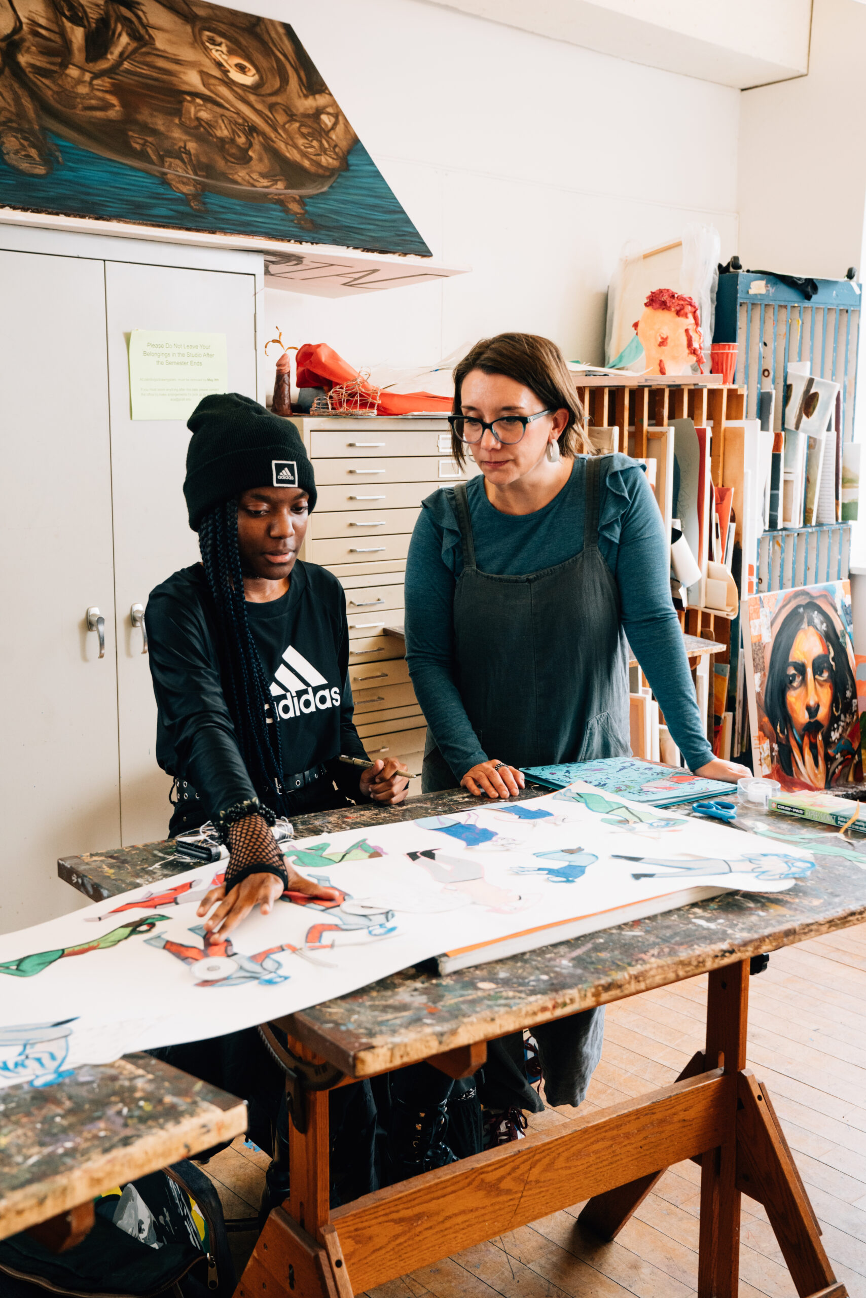 Two women are engaged in a creative discussion in an art studio. One is wearing a black beanie and a black Adidas sweatshirt, leaning over a large piece of artwork on the table. The other, wearing a blue top and gray overalls, looks attentively at the artwork and gestures with her hand. Various art supplies and colorful artwork are visible around them, highlighting a collaborative and artistic environment.