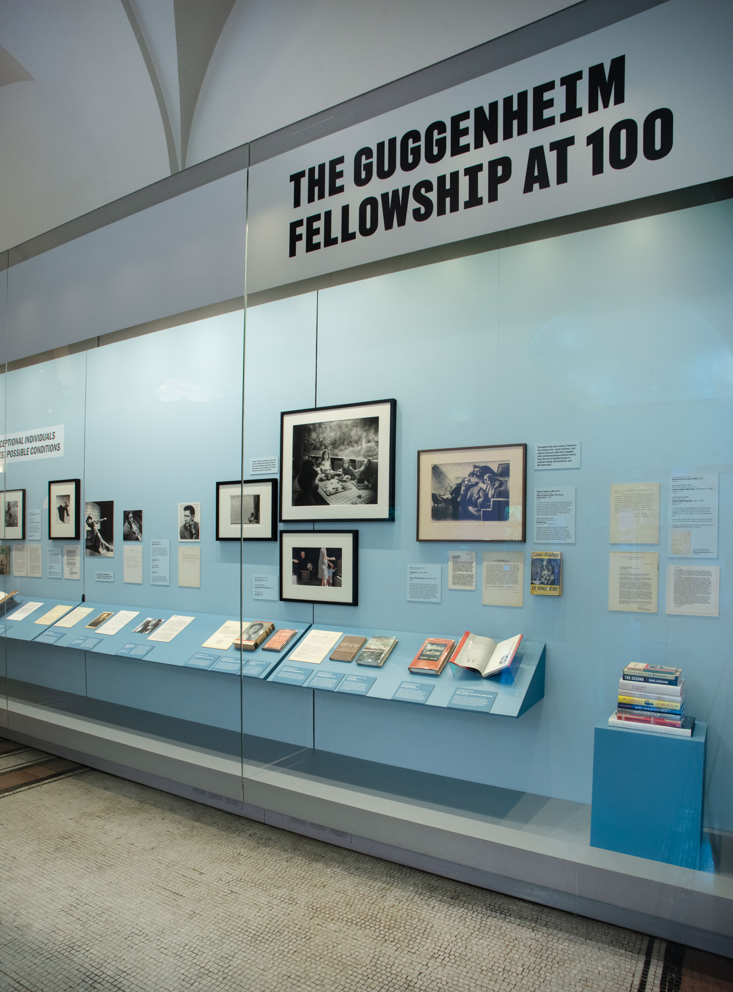 A museum display showcases "The Guggenheim Fellowship at 100." The exhibit includes framed photographs and various documents on a light blue background.