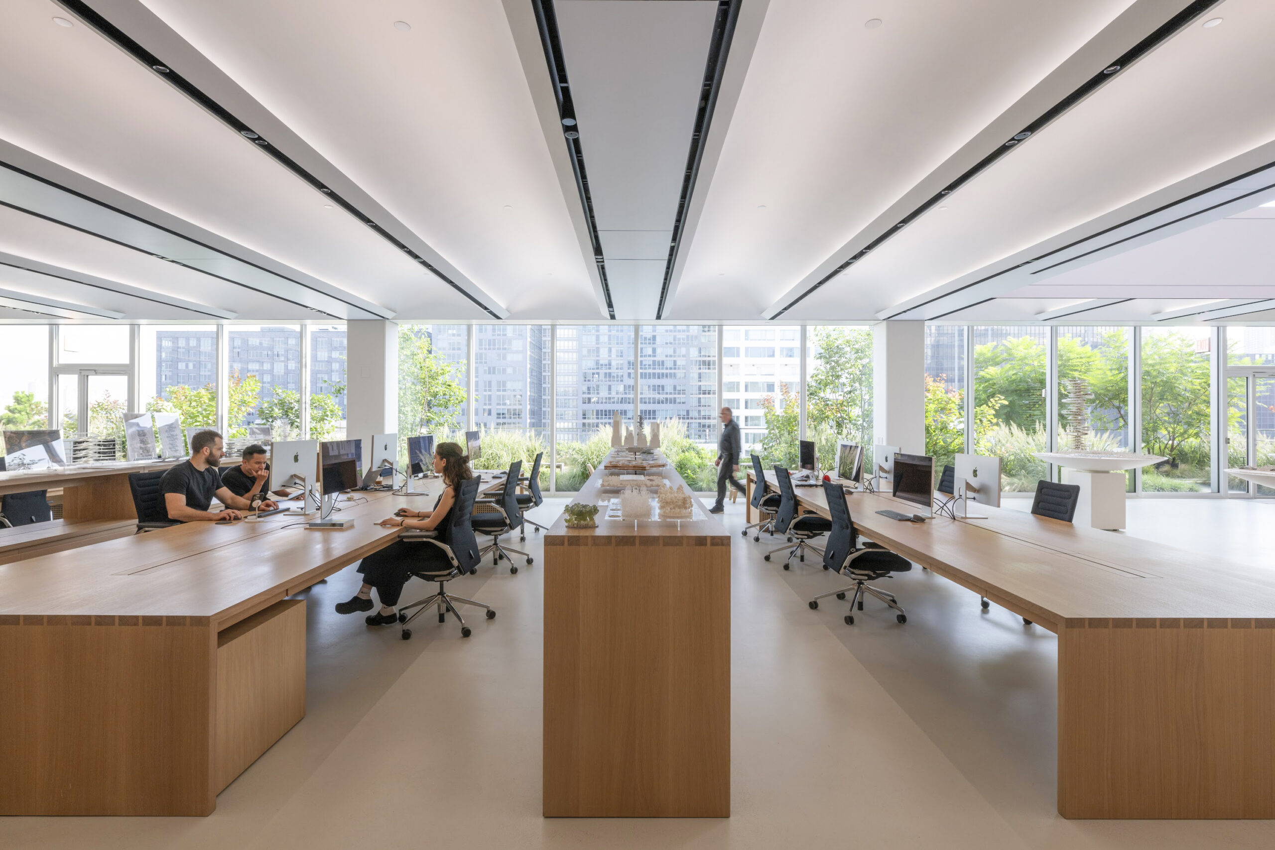 A modern office space with large windows allowing natural light to flood in. Several people work at long wooden tables with computers. A central wooden structure holds architectural models.