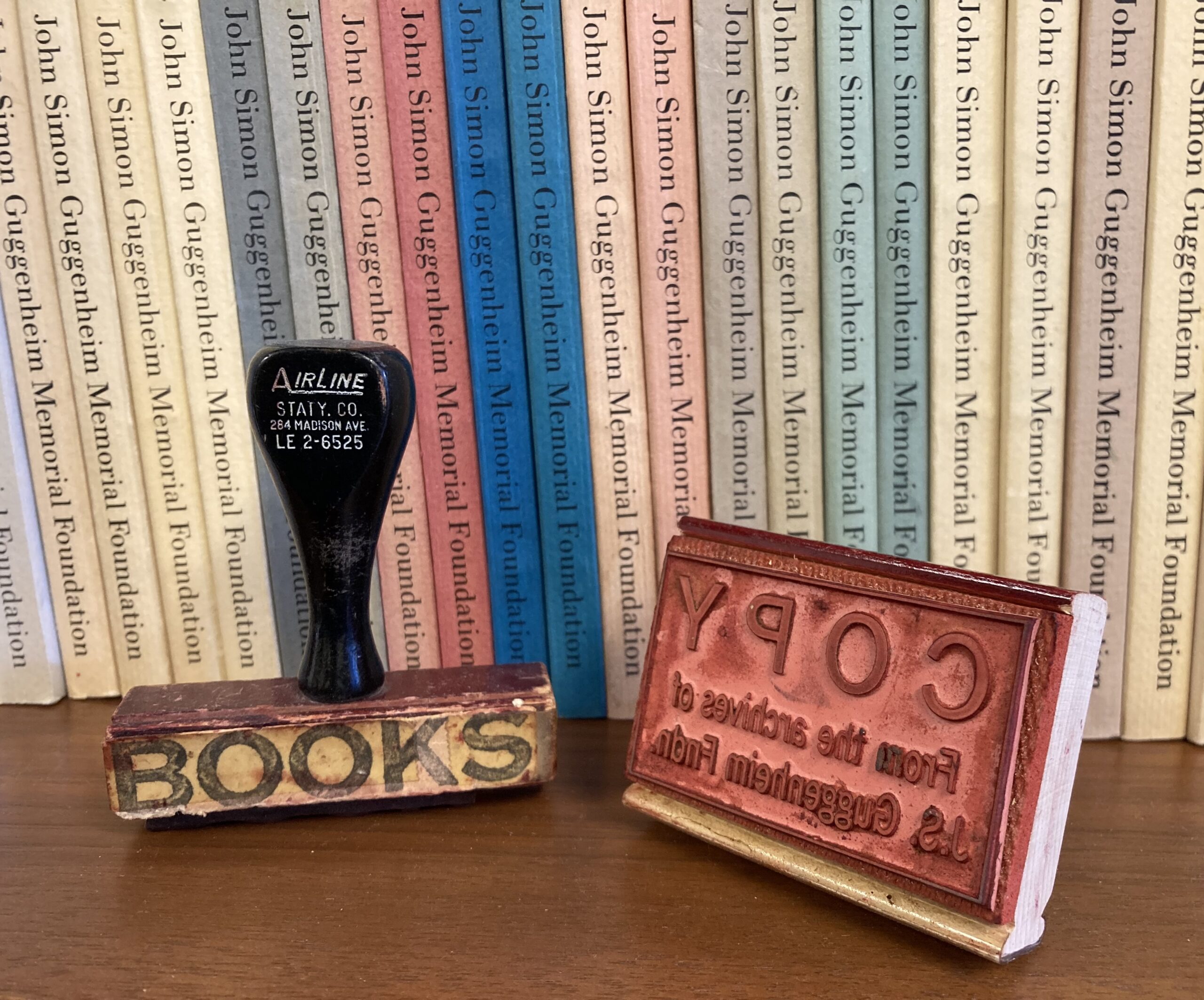 A large vintage stamp that says “Books” sits next to another stamp that reads “Copy from the archives of the J.S. Guggenheim Fndn.” They are placed before a row of books that read “John Simon Guggenheim Memorial Foundation” on each spine.