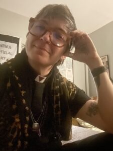 A person with short, tousled hair is sitting with a slight smile. They are wearing large, rectangular glasses and a black outfit with a patterned scarf. Various necklaces hang around their neck, and they have visible tattoos on their forearm. The background features a wall with posters and notes.