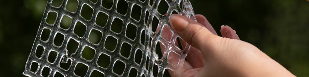 A close-up of a hand holding a transparent, flexible mesh panel with a grid pattern. The background is blurred greenery, suggesting an outdoor setting.