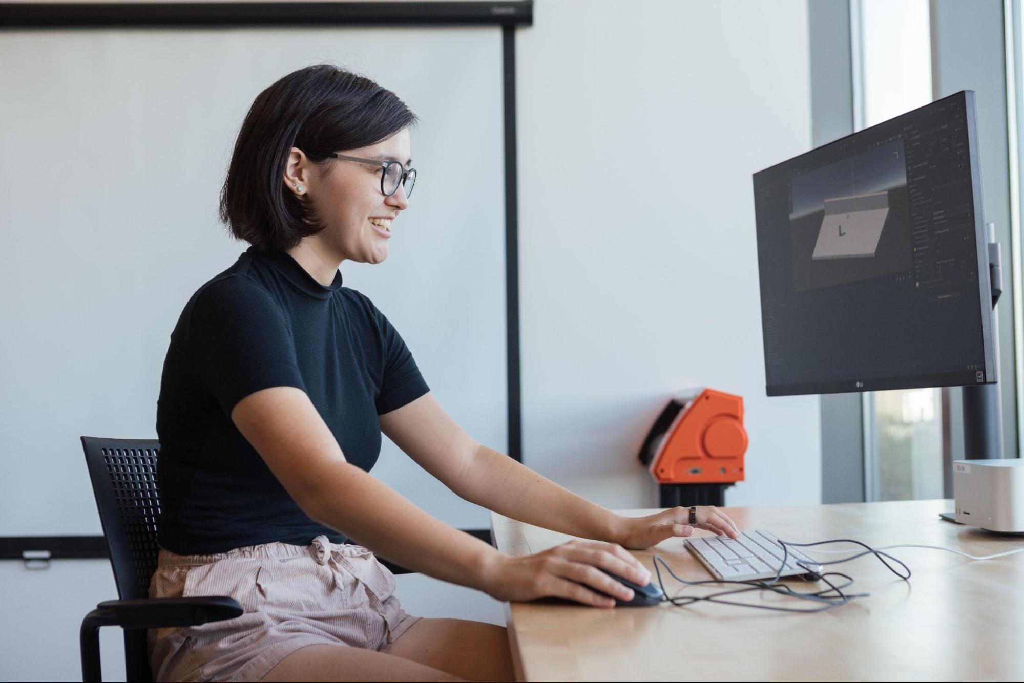 A person wearing glasses sits at a desk using a computer in a bright office setting. They hold a mouse with one hand and type with the other while looking at a large monitor displaying a 3D modeling or design interface. A keyboard and cables rest on the desk, and daylight enters from a nearby window.