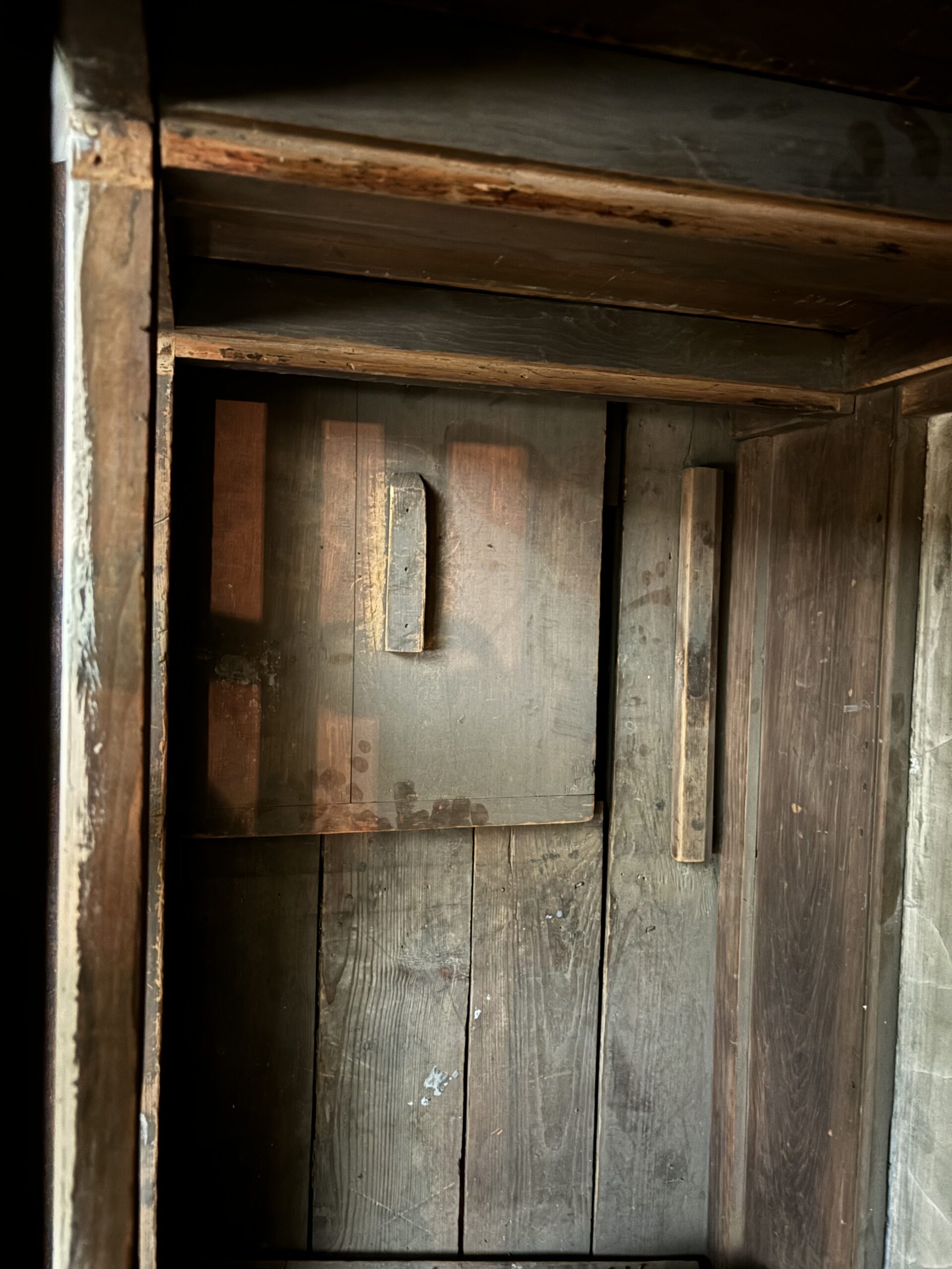 An interior view of a rustic wooden structure featuring dark, aged wood. There are two vertical wooden panels on the right and a rectangular inset on the left side of the wall. The floor is made of planks showing signs of wear, with a few scattered marks. Natural light illuminates the space, emphasizing the texture and grain of the wood.