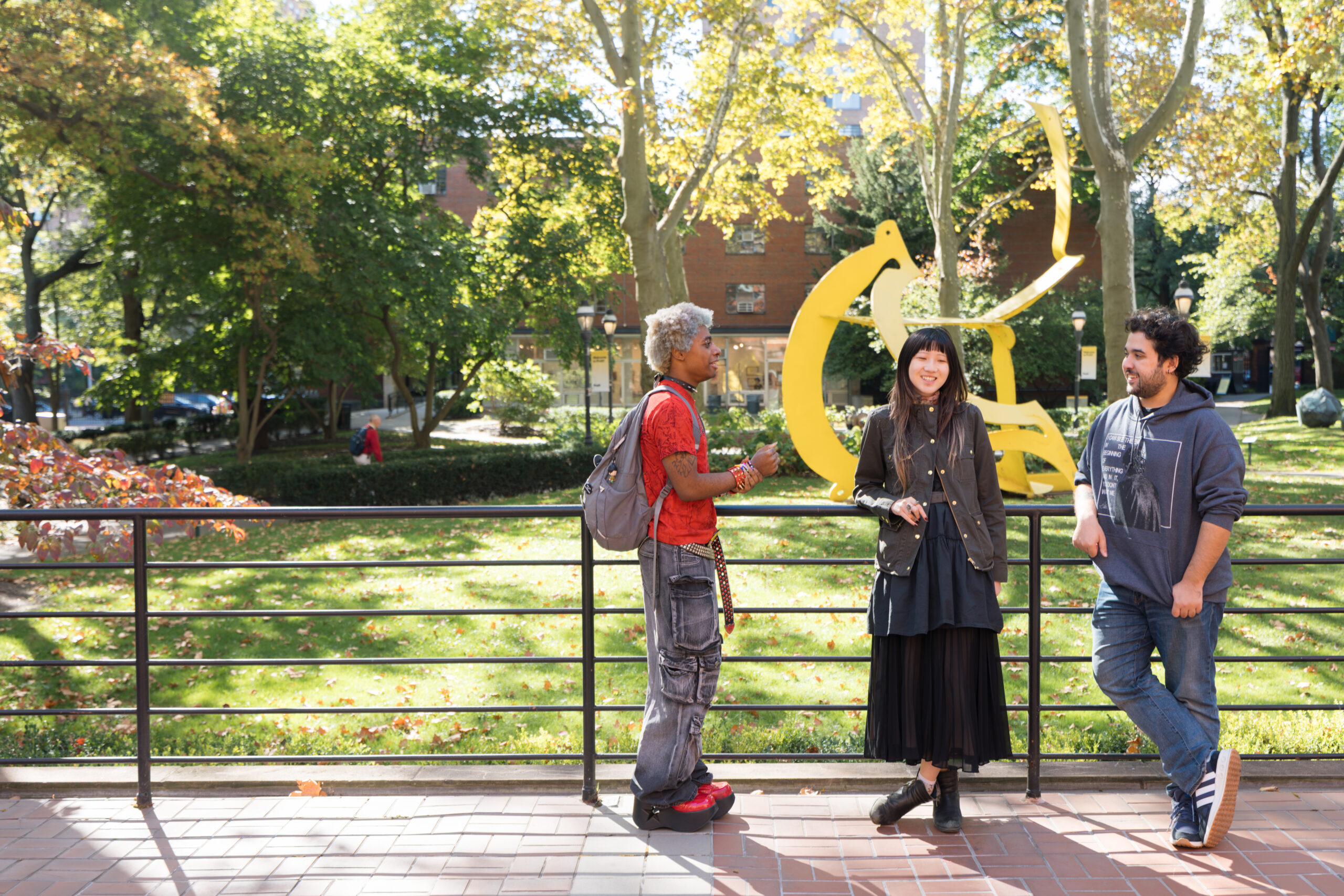 Three people stand together in a park setting with trees and a yellow abstract sculpture in the background. The person on the left has silver hair and wears a red shirt, gray pants, and red shoes. The person in the center, dressed in black, smiles while holding a phone. On the right, a man in a gray hoodie and denim jeans leans casually against the railing. The ground is covered in fallen leaves, indicating autumn.