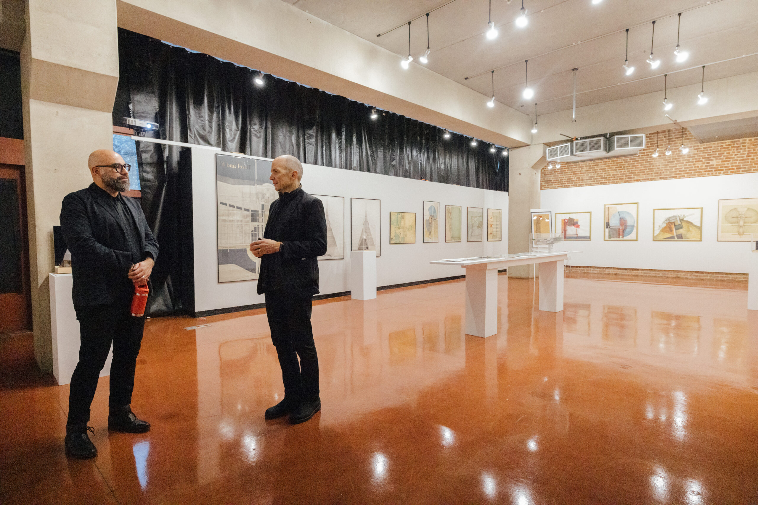 Two men stand conversing in a contemporary art gallery. Both wear dark clothing. One man with glasses holds a red bottle at his side, while the other gestures with his hands as they speak. Framed artworks hang along the white walls, and a central display table stands in the middle of the room. The gallery has polished brown floors, exposed brick accents, and ceiling-mounted track lighting.