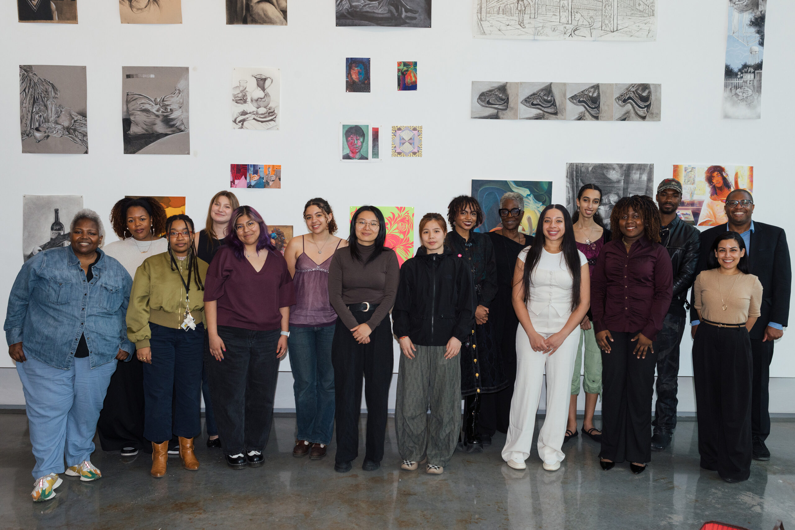 A diverse group of 17 individuals poses together in front of an art wall displaying various drawings and paintings. They are smiling and dressed in a mix of casual and formal attire. The background features a variety of artworks, including portraits and still life, giving a creative atmosphere to the gathering.
