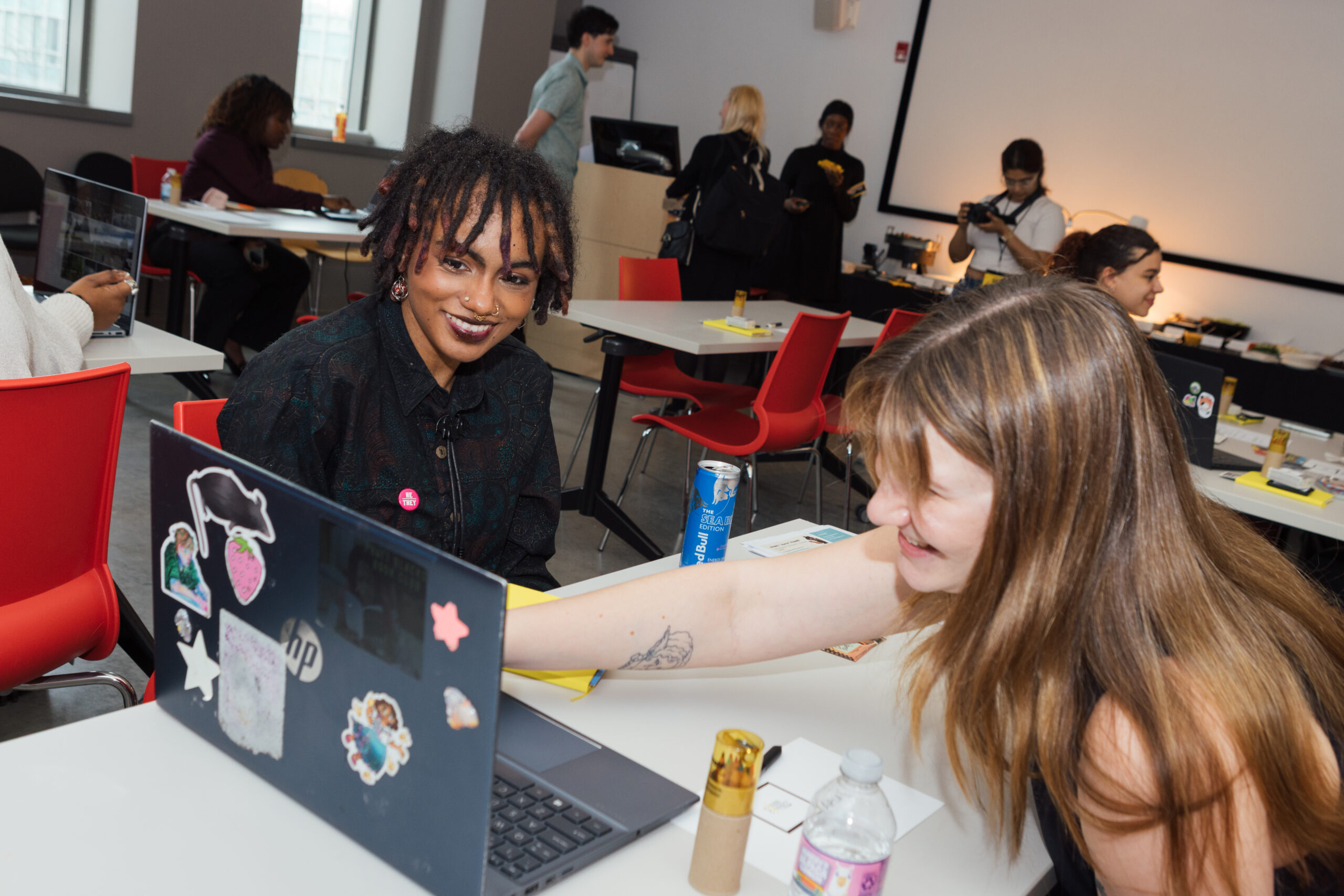 Two young women are engaged in a discussion at a table with laptops. One woman, with dark, curly hair, smiles while wearing a button and dark clothing. The other woman, with long, light brown hair, leans in to point at the laptop screen, looking excited. The background shows other people working and a projector screen. The setting appears to be a collaborative workspace or event.