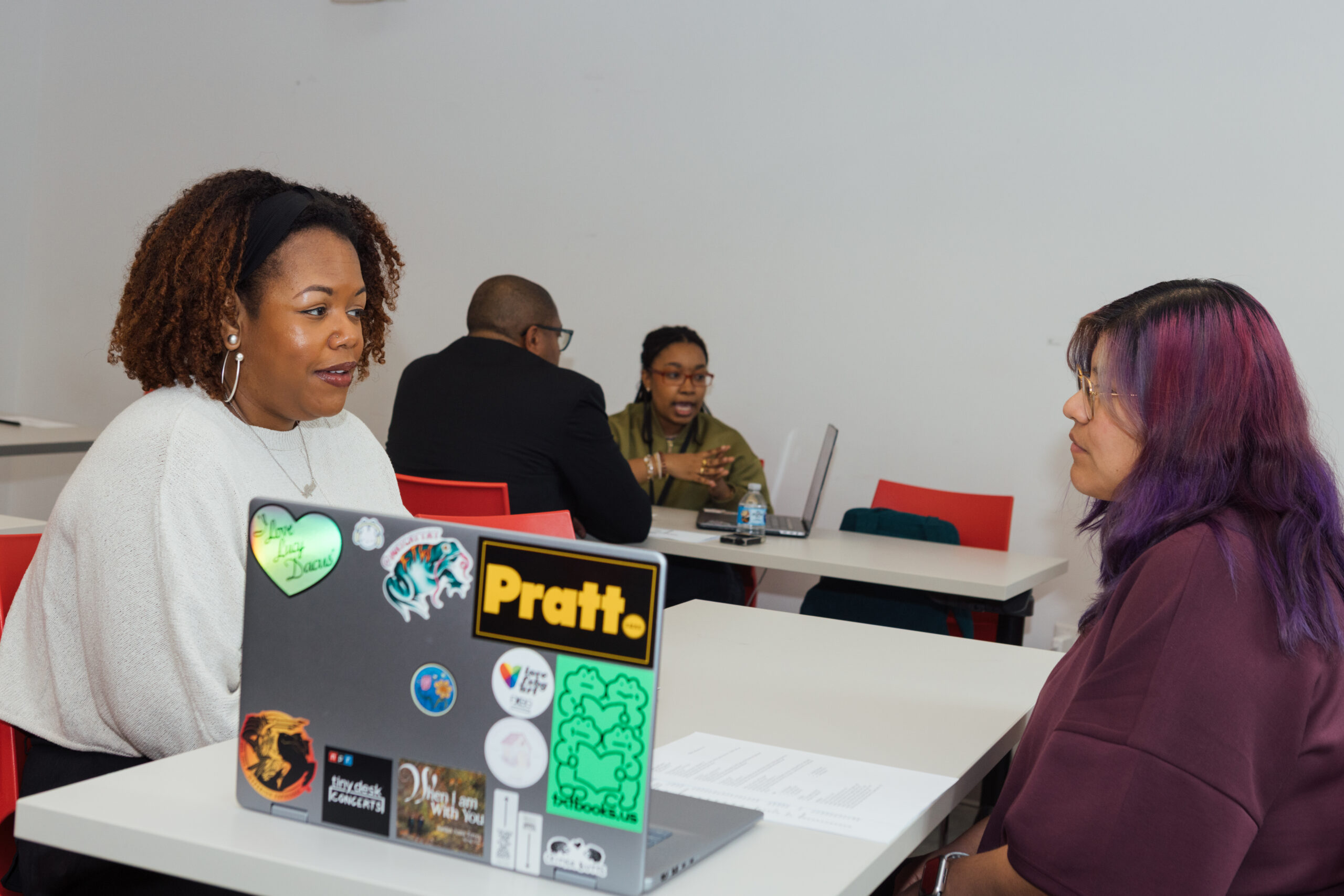 Two women are seated at a table engaged in conversation. The woman on the left has curly hair and is wearing a light sweater, while the woman on the right has long hair with purple highlights and wears glasses. In the background, a man in a suit and another woman are also having a discussion. A laptop covered in stickers sits in front of the first woman. The setting is a bright, modern room with red chairs.