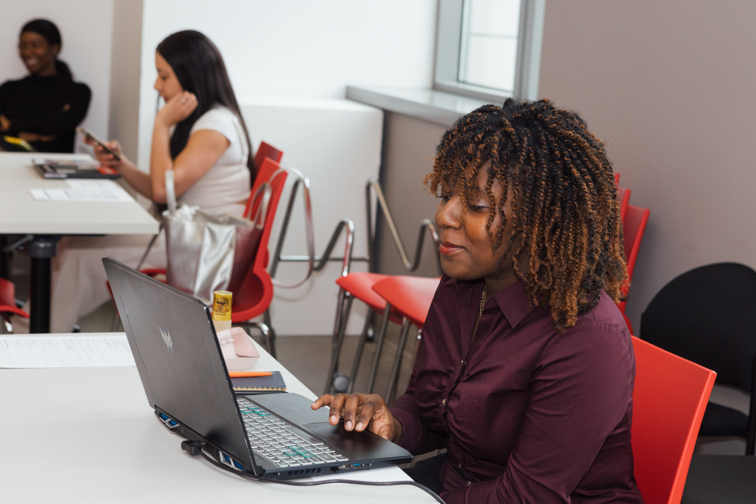 A woman with curly hair is seated at a table, using a laptop. She is wearing a burgundy shirt and appears focused on her work. In the background, a second woman is sitting and looking at her phone, while another woman is partially visible. Red chairs and a modern interior can be seen in the setting.