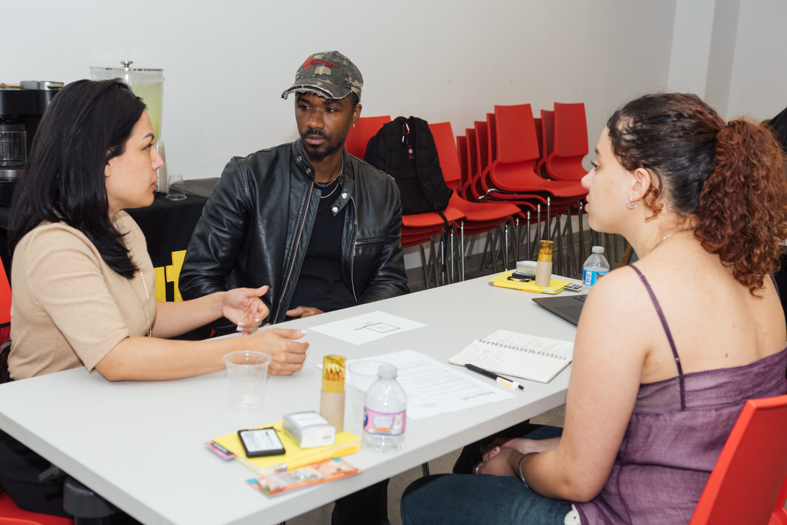Three people are seated around a table engaged in conversation. One woman in a beige top gestures while speaking, while a man in a black leather jacket listens attentively. The third person, a woman in a purple top, sits with her hands clasped, showing interest. In the background, there are red chairs stacked. Items on the table include a water bottle, notebooks, and a cup.