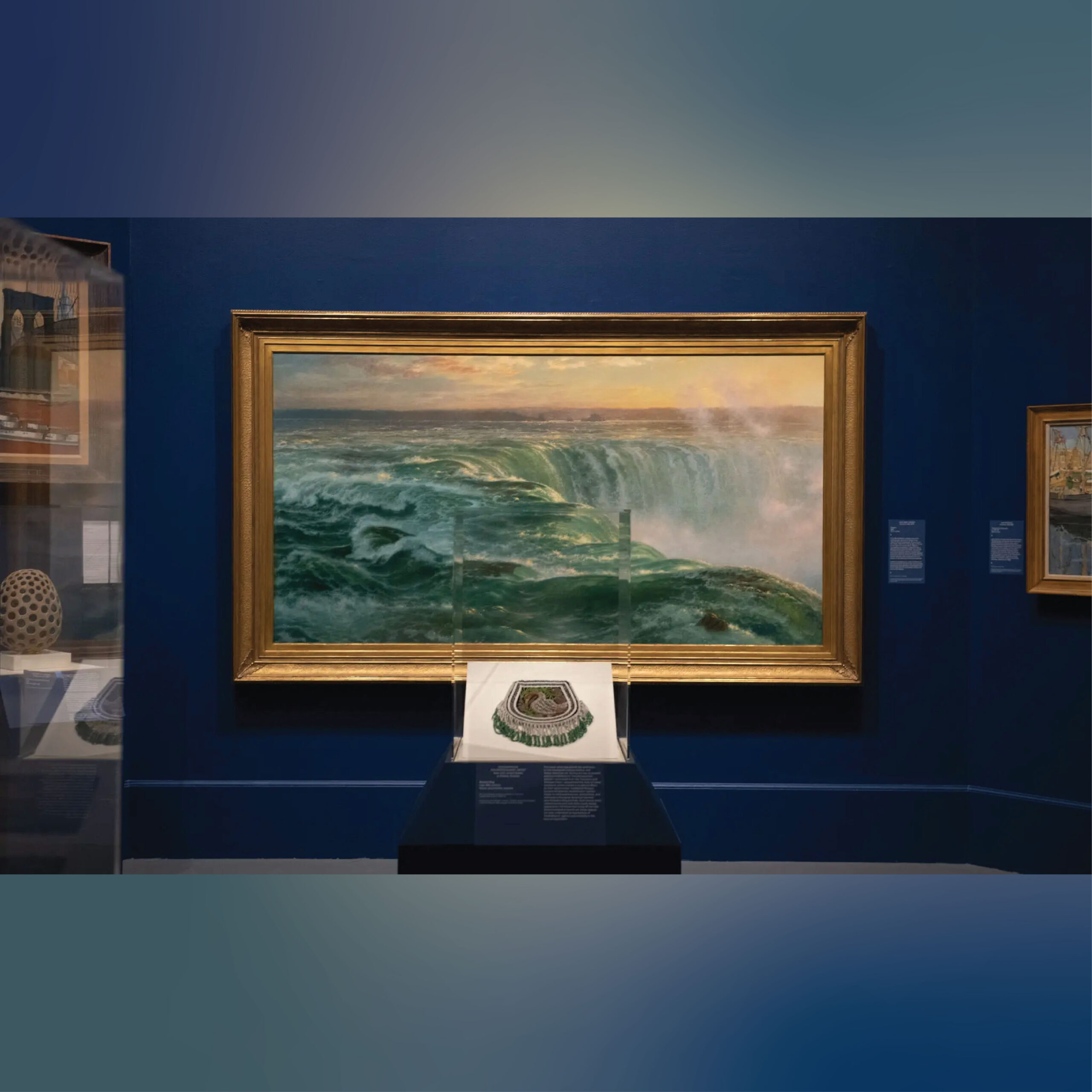 Large framed seascape painting displayed on a deep blue gallery wall. The painting shows powerful green waves cresting over the edge of a waterfall at sunset, with mist rising in the distance. A small object is mounted on a pedestal directly below the painting with a descriptive label. Additional framed artworks and wall labels are partially visible to the right, and a glass display case appears to the left.