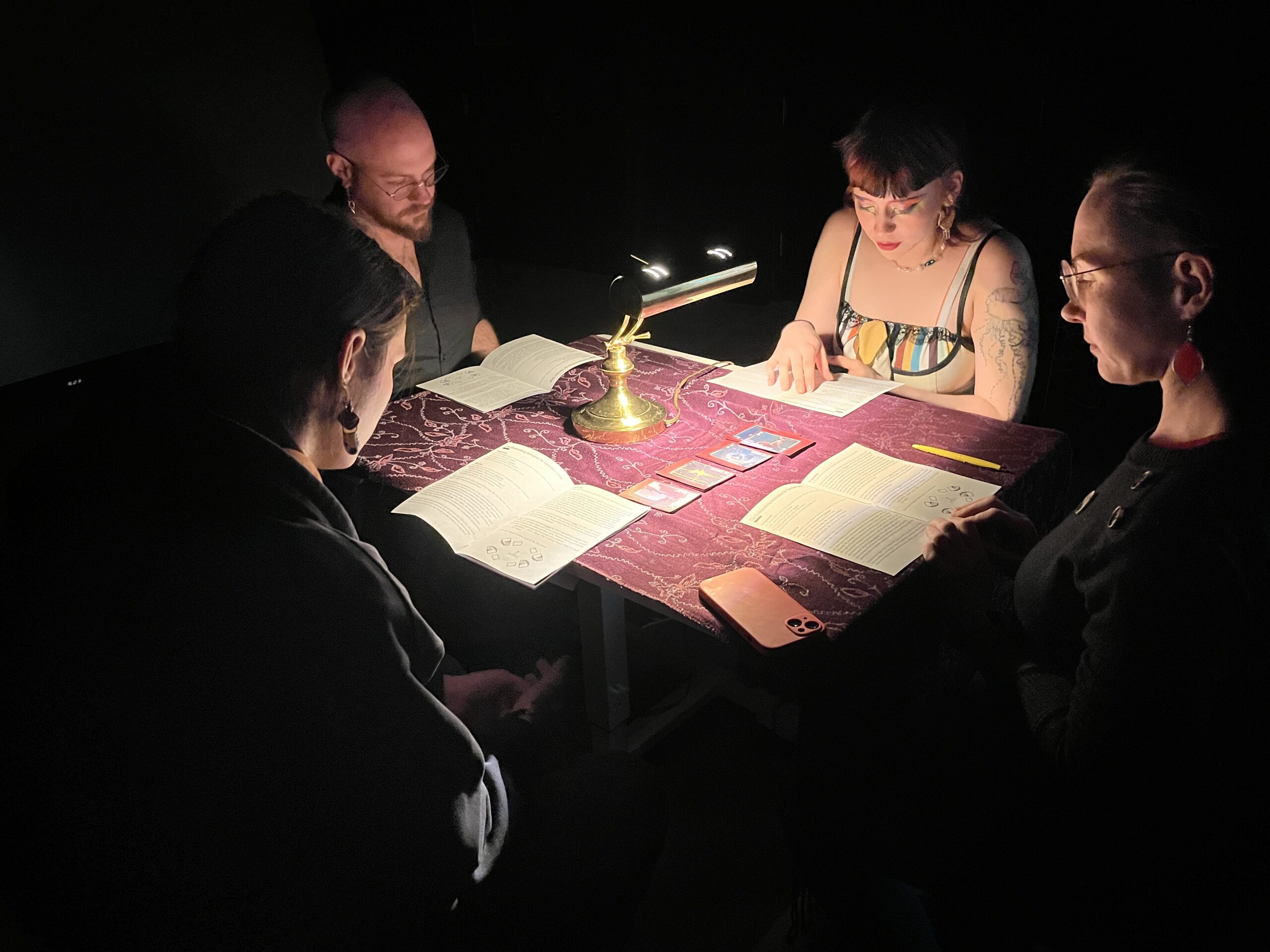 A group of four people sits around a table covered with a patterned cloth, illuminated by a brass lamp. They are engaged in a card game or tabletop role-playing game, with rulebooks and colorful cards spread out before them. Each person is focused on the game, with varying expressions of concentration. The scene is dimly lit, creating an intimate atmosphere.