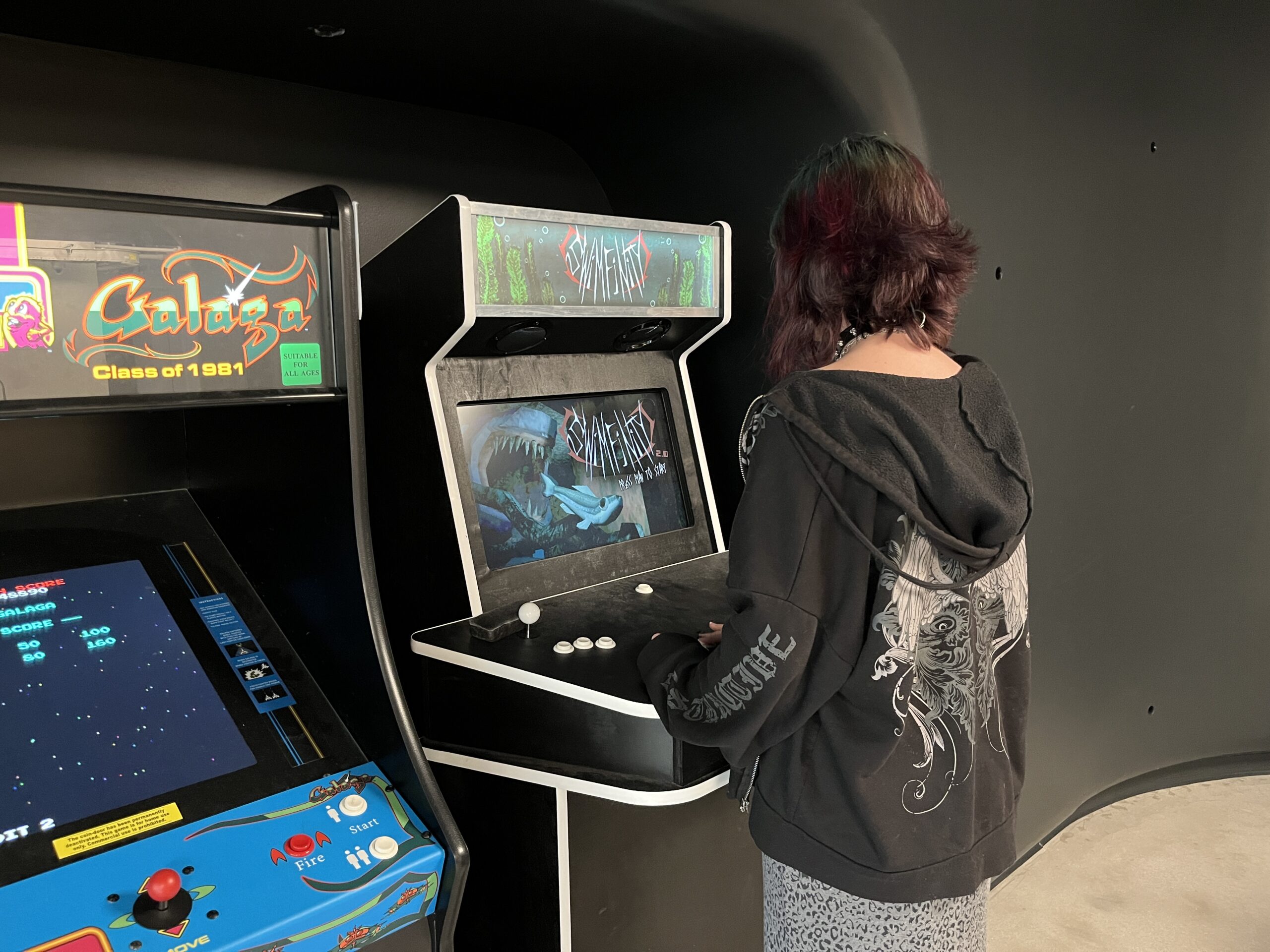 A person with multicolored hair stands in front of two arcade machines. They are facing a machine displaying a game called "Mantis," which features an image of a shark. Another machine, labeled "Galaga," is visible to the left. The setting has a dark background, and the person is wearing a black hoodie with a graphic design on the back. The floor is smooth and appears to be made of concrete.