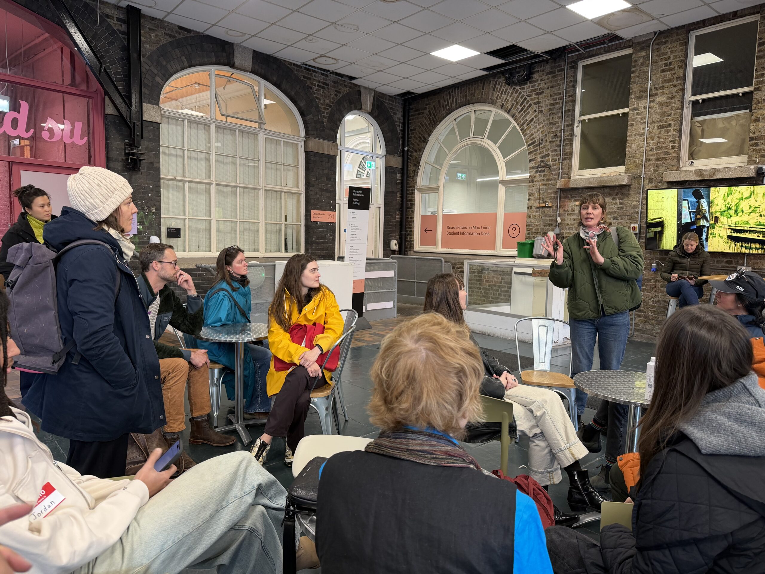 A group of people is gathered in a casual indoor setting, listening to a person speaking animatedly. They wear a green jacket and gesture with their hands while addressing the audience. The attendees, dressed in various colorful jackets, appear engaged and attentive. Behind them, there is a large screen displaying an image. The room features a mix of stone walls and modern furniture.