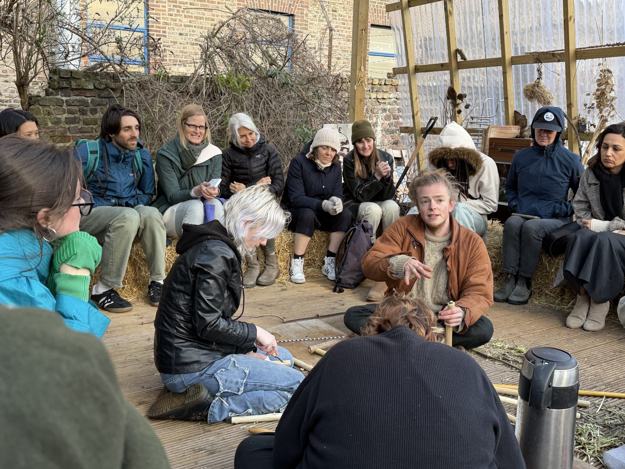A diverse group of people sits in a semi-circle in a rustic, greenhouse-like setting. In the foreground, a person with light-colored hair demonstrates a technique using a tool while others observe intently. Some individuals are wearing warm clothing and gloves, suggesting a cold environment. The atmosphere appears focused and engaged, with various plants and natural materials visible around the space.