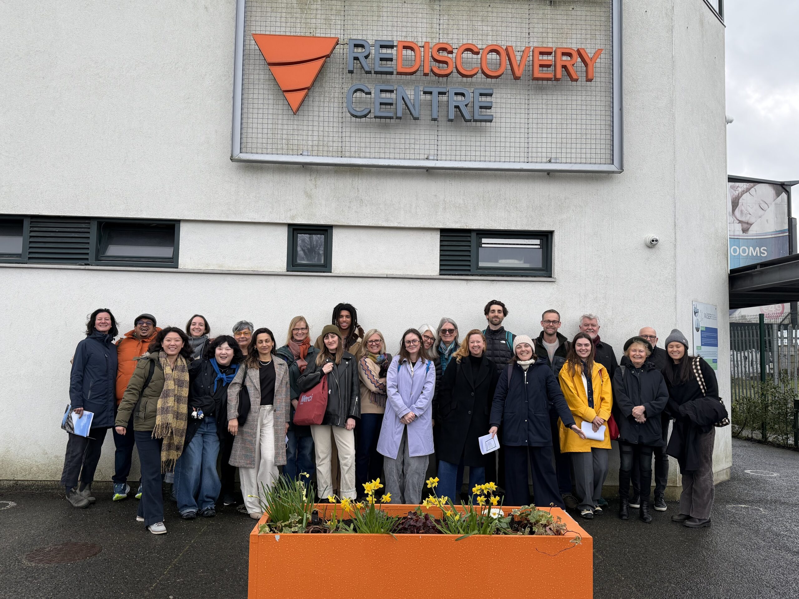 A diverse group of approximately 25 people stands in front of the Rediscovery Centre, an industrial-style building with a sign above. The people are smiling and appear to be posing for a photo. In the foreground, there is a bright orange planter with blooming flowers. The sky is overcast, indicating a cloudy day.