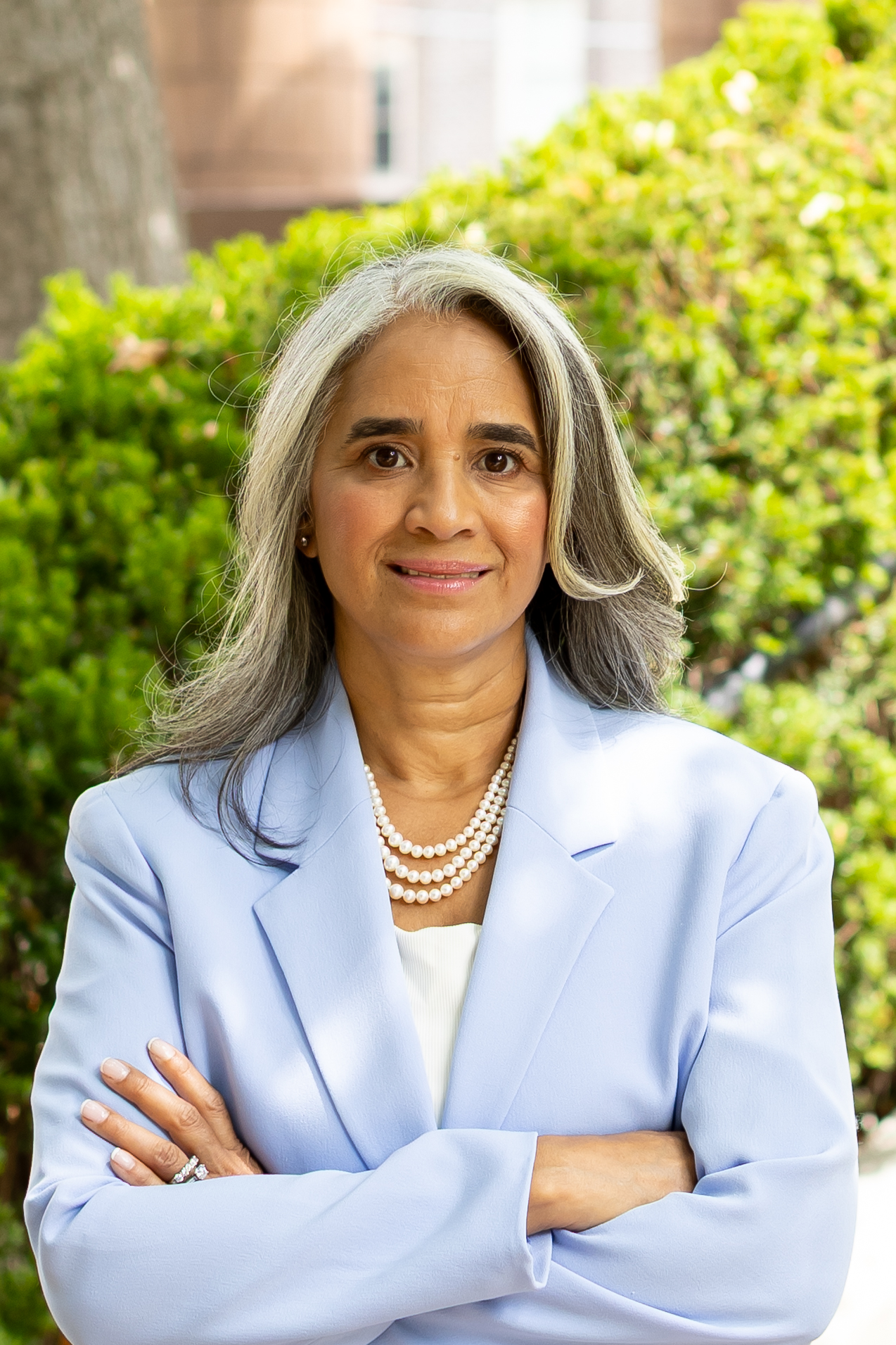 A woman with long, wavy gray hair stands outdoors, wearing a light blue blazer and a pearl necklace. She smiles softly with her arms crossed in front of her, surrounded by greenery in the background. The photo has a bright, natural light setting.
