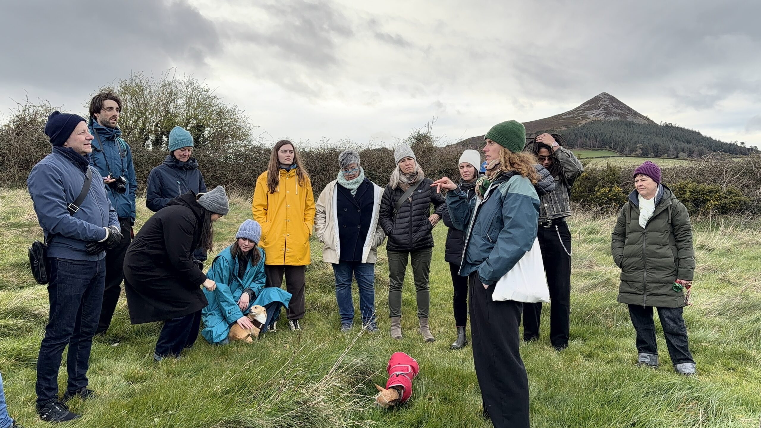 A group of people is gathered in a grassy field, listening attentively to a person speaking. Some individuals are sitting while others are standing, wearing winter clothing like jackets and hats. Two small dogs are present, one wearing a red coat. In the background, a small mountain and cloudy sky are visible.