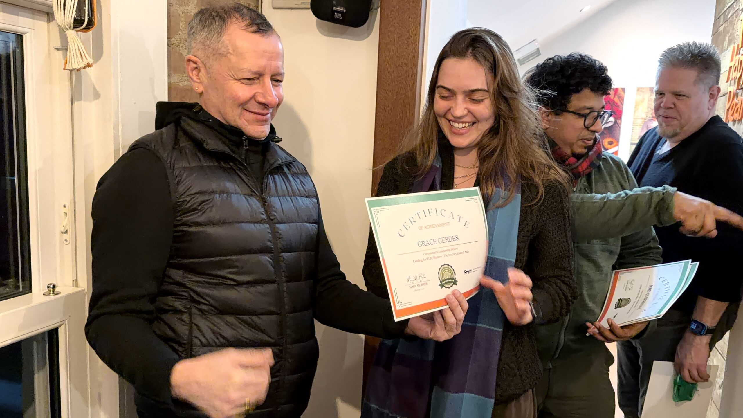 Two people are smiling and holding a "Certificate of Achievement" as they engage in a celebratory moment. A man in a black vest stands beside a woman who has long hair and is dressed in black and a blue scarf. In the background, a few others can be seen, including a person wearing glasses and a scarf.