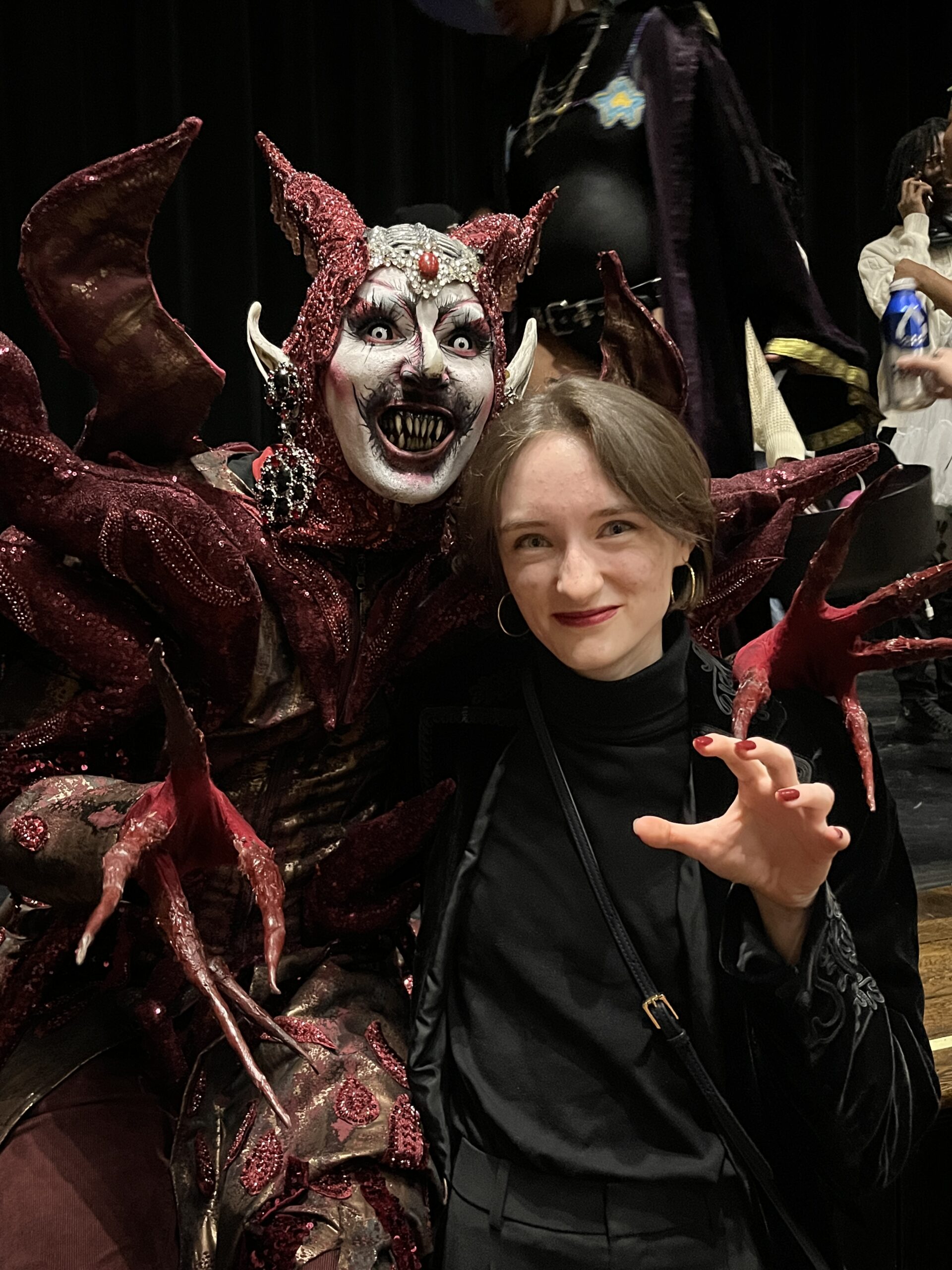 A person in elaborate, dark and glittery fantasy makeup and costume poses next to another individual, who is wearing a black outfit and playfully mimicking a claw-like gesture. Both are smiling, with the fantastical figure displaying exaggerated facial features, including sharp teeth and horns, while the background features blurred figures in colorful outfits.