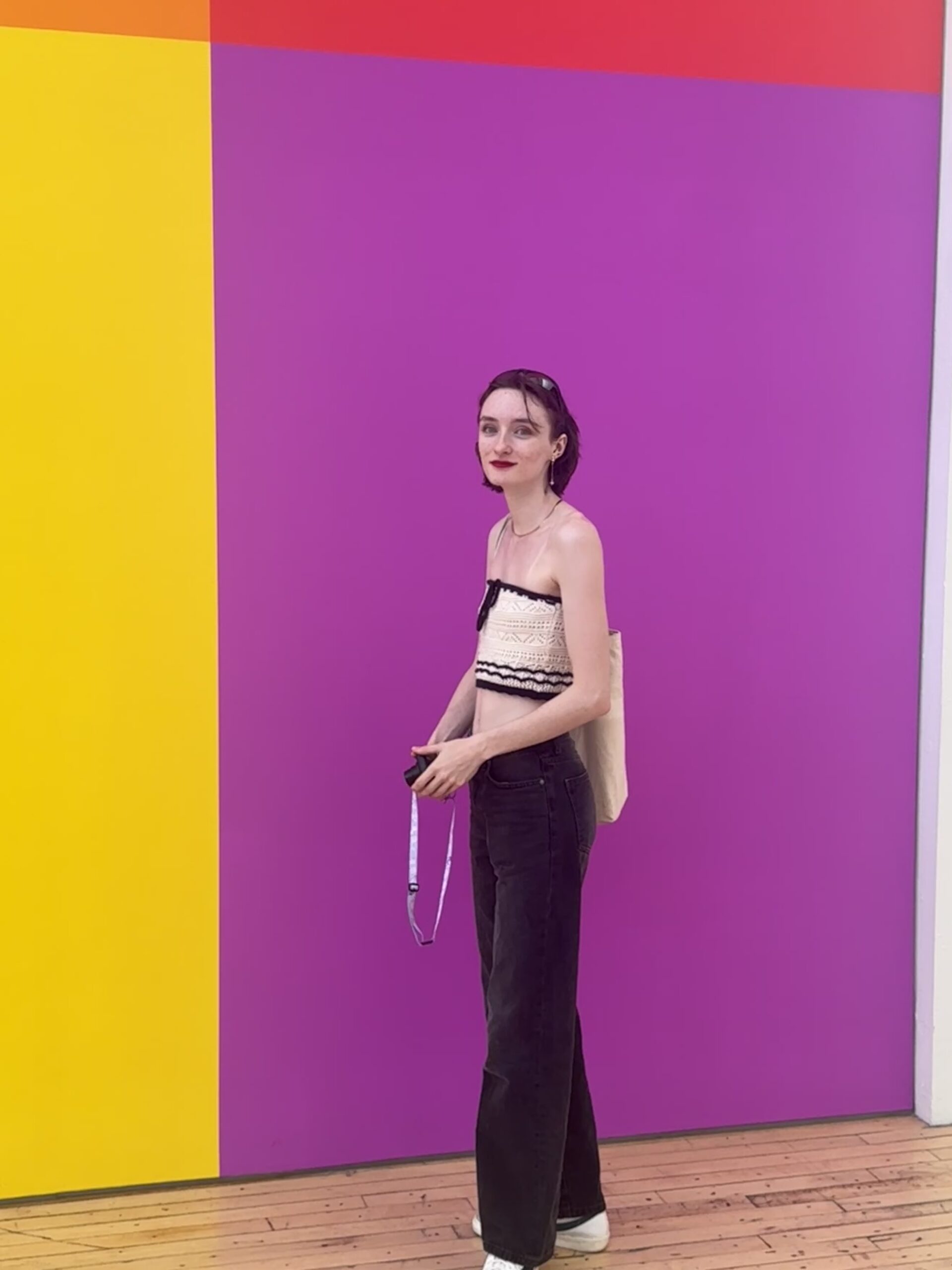 A person stands against a bright, colorful backdrop featuring a vertical split of yellow and purple with a red section at the top. They are wearing a cropped, strapless top with a black design and black jeans, holding a camera in one hand. Their hair is short and reddish-brown, and they have a light makeup look, including red lipstick. The floor is wooden and adds a warm tone to the scene.