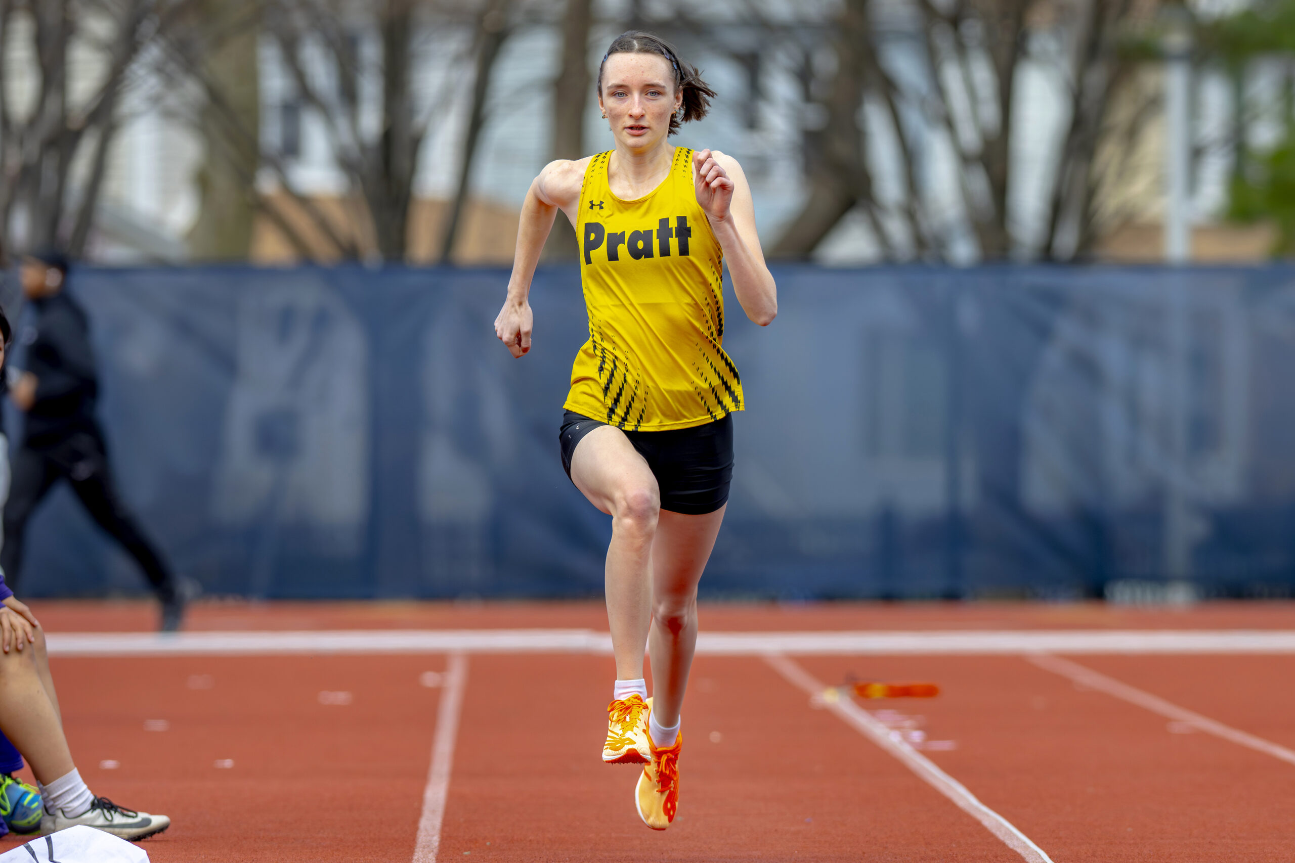 An athlete is sprinting on a track, wearing a yellow tank top with "Pratt" printed on it and black shorts. Her running form is dynamic, and she has orange running shoes. In the background, there are blurred figures of other athletes and trees, suggesting an outdoor track event. The surface of the track appears red, and there are markers visible nearby.