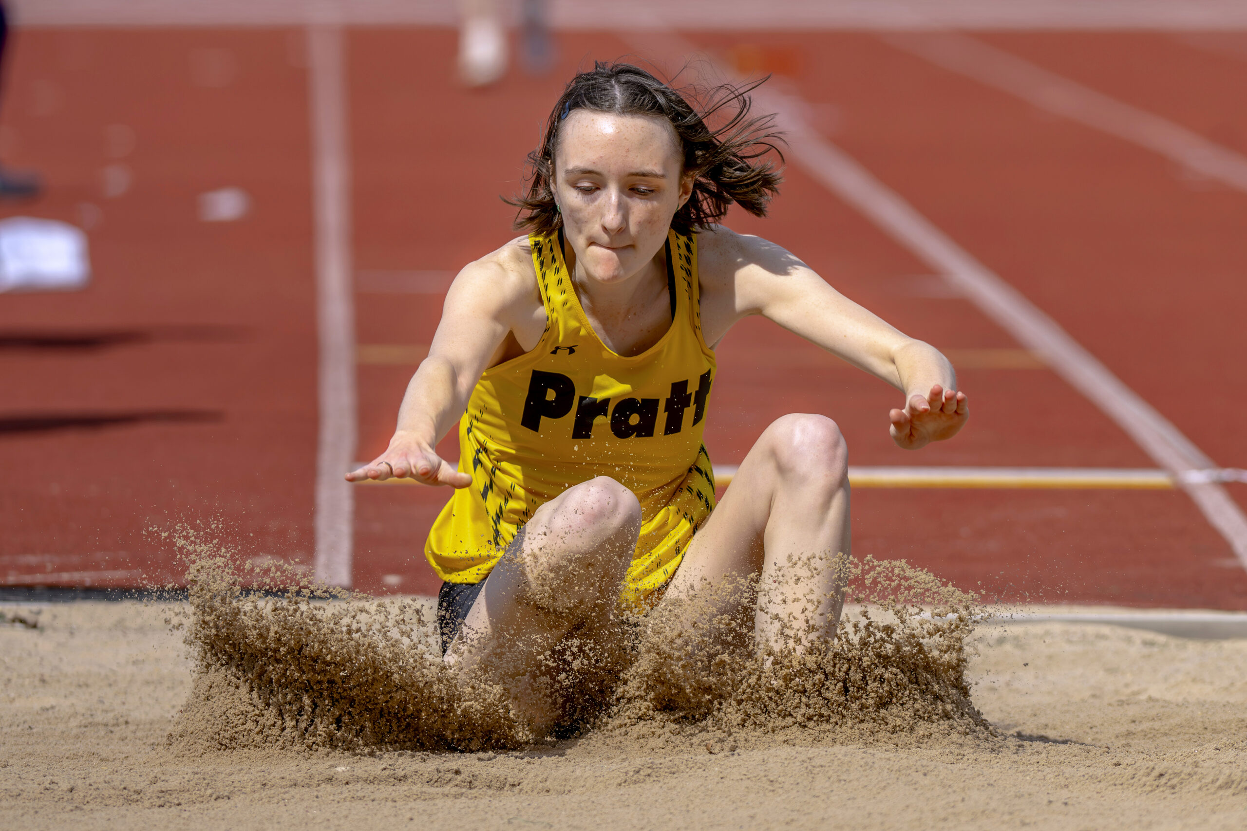 An athlete in a yellow and black uniform is mid-jump in a sand pit during a track event. She is positioned in the air with her arms extended forward, and sand is being displaced behind her as she lands. The background features a red track surface and a blurred crowd or view of the field.