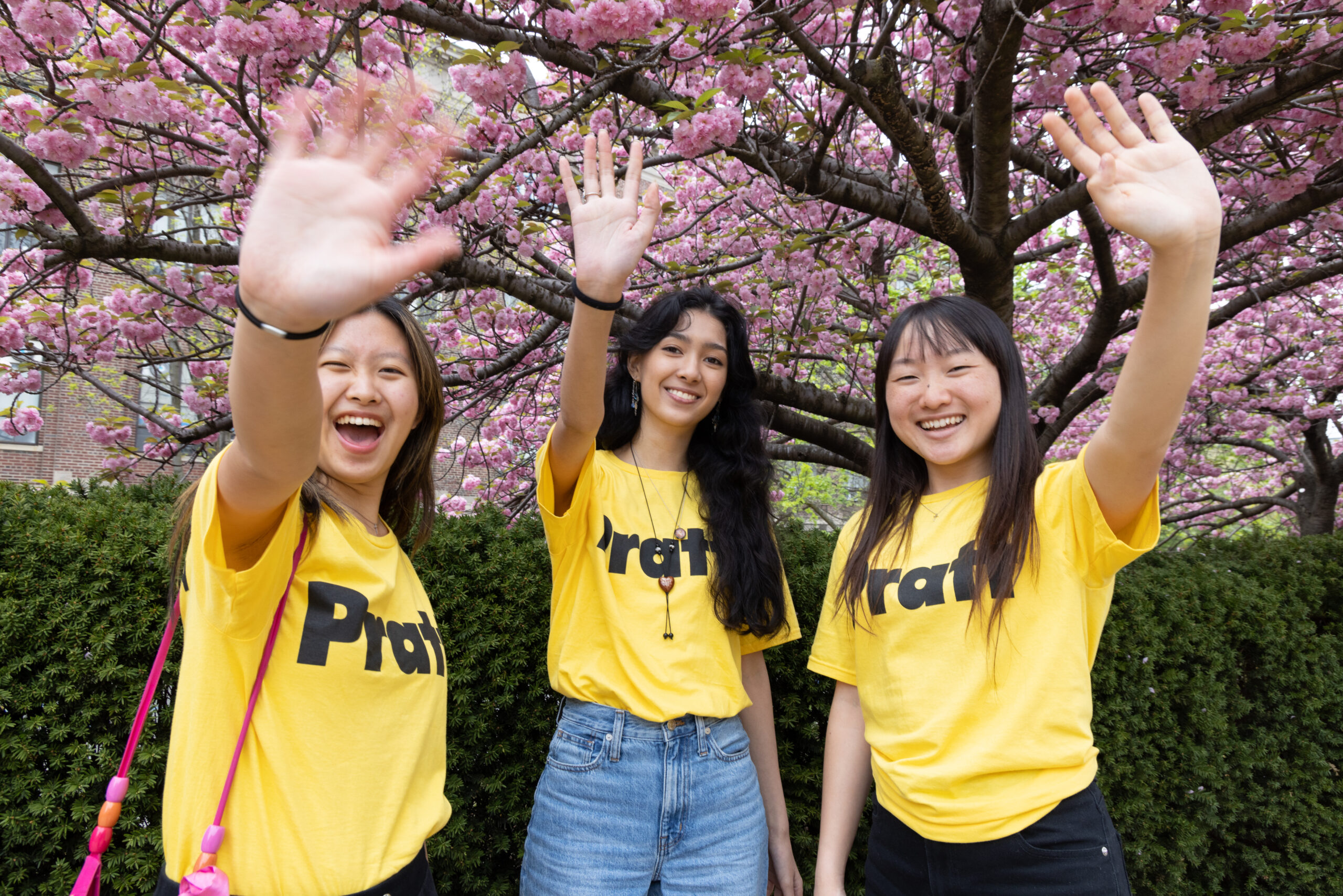 Three young women are smiling and waving at the camera, standing in front of a flowering pink cherry blossom tree. They are wearing matching yellow t-shirts with the word "Pratt" printed in large black letters. The woman in the center has long, wavy hair, while the women on either side have straight hair. They appear joyful and engaged in a cheerful moment.