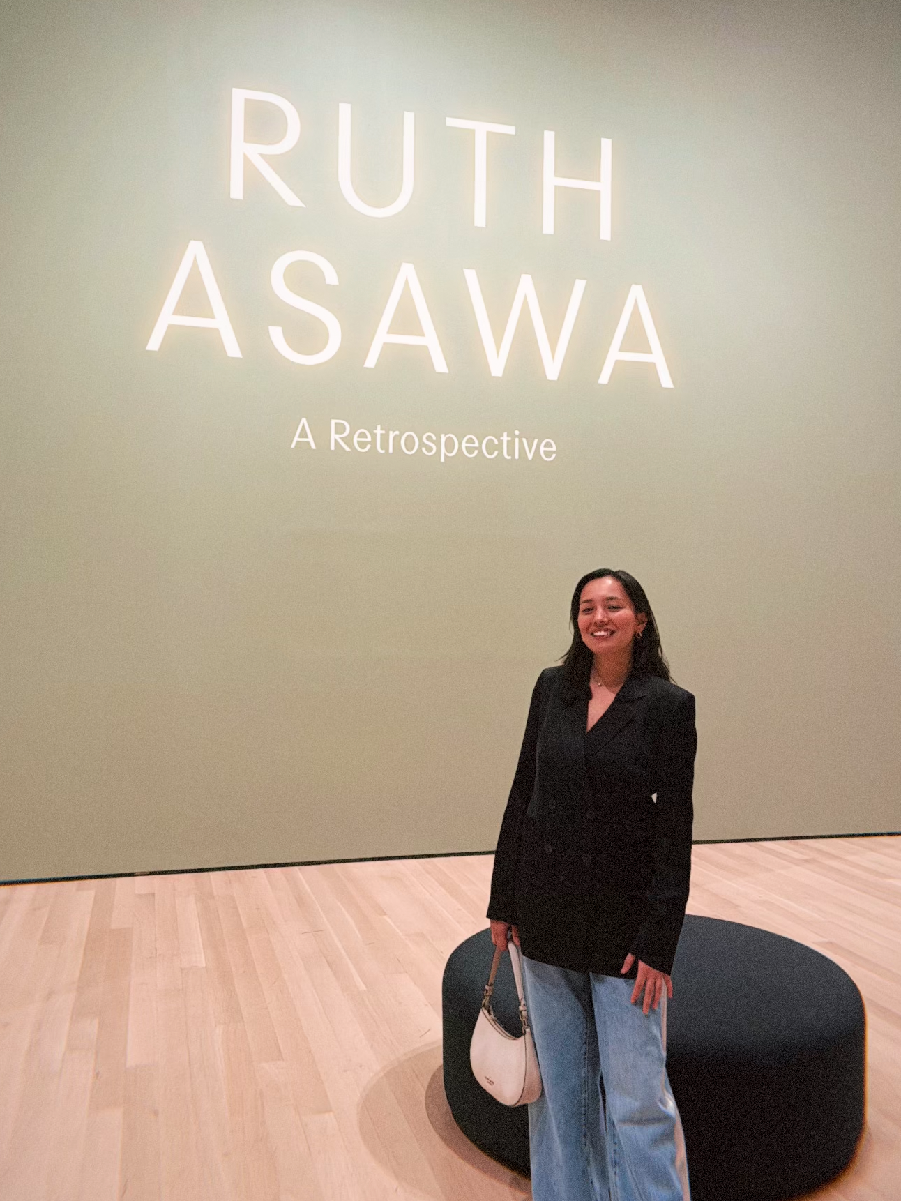A woman stands smiling in front of a large exhibition wall that reads "RUTH ASAWA A Retrospective." She wears a black blazer and light blue wide-leg jeans, holding a small white handbag. A black circular seating area is positioned in front of her, and the floor is made of light wood.