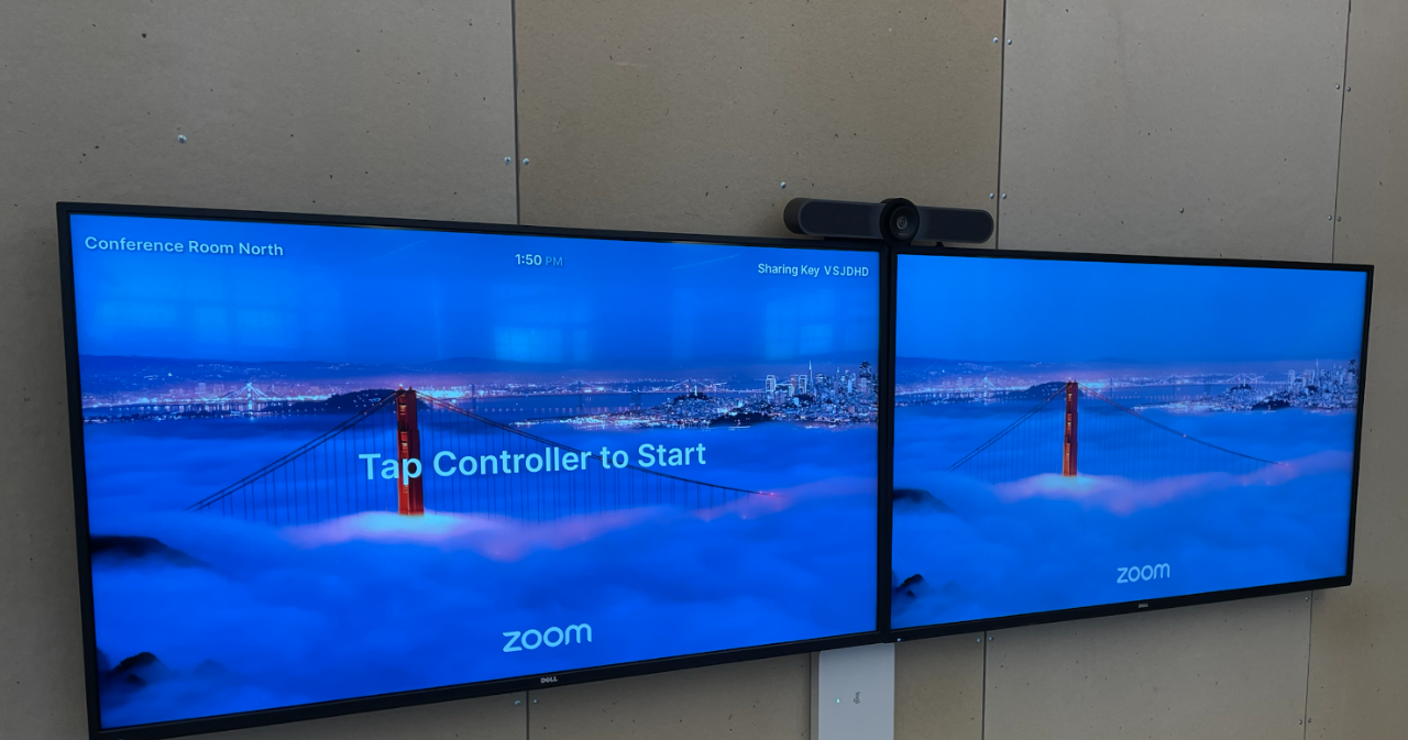 Two screens are displayed in a conference room setting, showing a scenic view of the Golden Gate Bridge emerging from fog at dusk. The left screen indicates "Conference Room North" and the time "1:50 PM," while the right screen prompts users to "Tap Controller to Start" with a sharing key displayed. A webcam is mounted above the screens, and the Zoom logo is visible at the bottom of each screen.