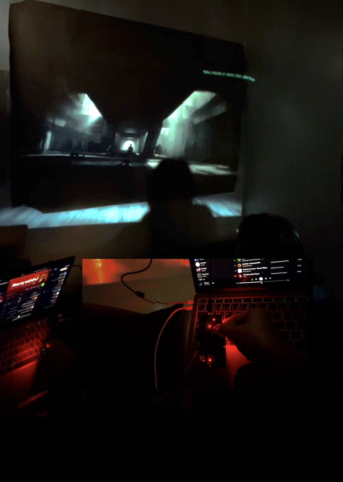 The image showcases a dimly lit room where a large screen displays a video game scene with dark, atmospheric lighting. In the foreground, a laptop keyboard is visible, illuminated in red light. A hand is adjusting a device connected to the laptop. The laptop screen shows a music streaming application, indicating it might be playing background music while gaming.