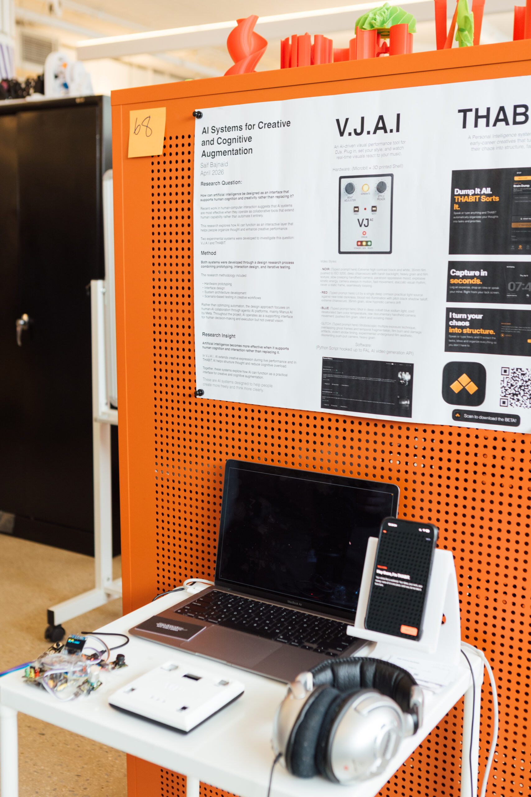 Exhibition display featuring a poster titled “AI Systems for Creative and Cognitive Augmentation” mounted on an orange perforated panel. Below the poster is a table with a laptop, smartphone on a stand, electronic components, cables, and over-ear headphones, indicating an interactive technology or research project setup.