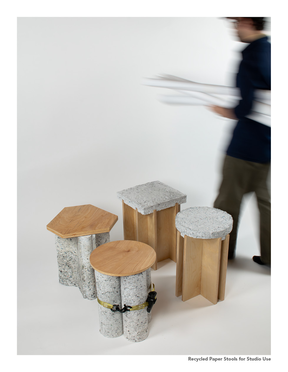 Four stools made from recycled materials are arranged on a white background. Two stools have wooden tops, one is hexagonal, and the other is circular. Two additional stools feature vertical cylindrical sections made from recycled paper, one of which is secured with a strap. A person in motion is blurred in the background, carrying sheets of paper.