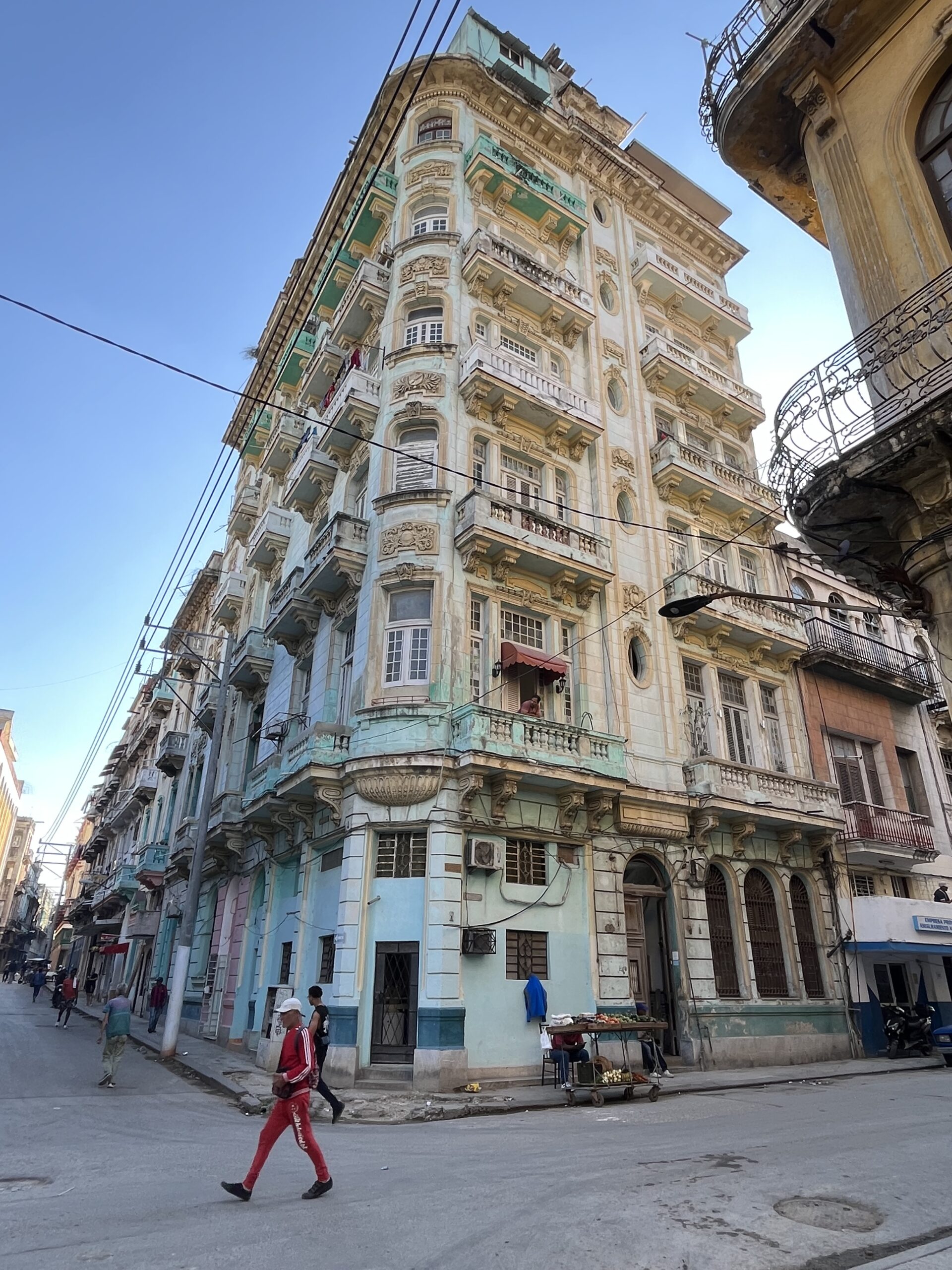 A tall, ornate building with a mix of colors, including pale blue and cream, stands at the corner of a street. The structure features intricate architectural details, balconies, and several windows adorned with shutters. Pedestrians are seen walking along the street, while a small vendor's cart displaying items is nearby. Power lines crisscross the scene under a clear blue sky.